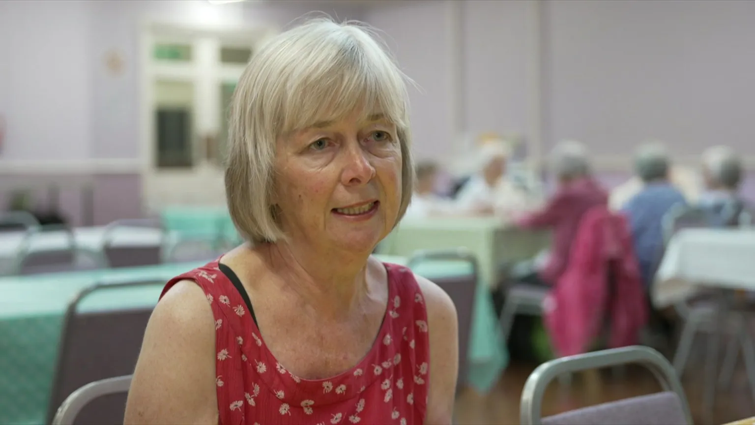 A woman with blonde hair in a bob sits in a community cafe with others in the background. She wears a red dress with floral print and has a serious expression on her face.
