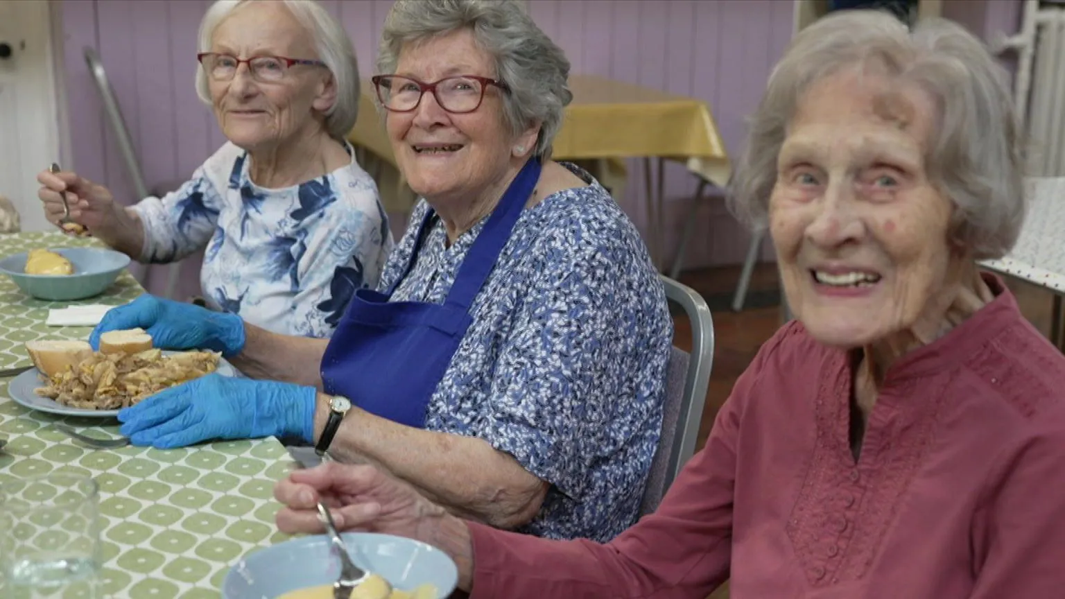 Three women sit next to each other at a table in a cafe. The woman on the far left is wearing glasses and a blue and white blouse. The woman in the middle is also wearing glasses. She is dressed in a navy blue apron on top of a blue and white floral dress. The woman on the right is wearing a mauve blouse. Two women are eating from a blue crockery bowl and the woman in the middle has a plate in front of her. 
