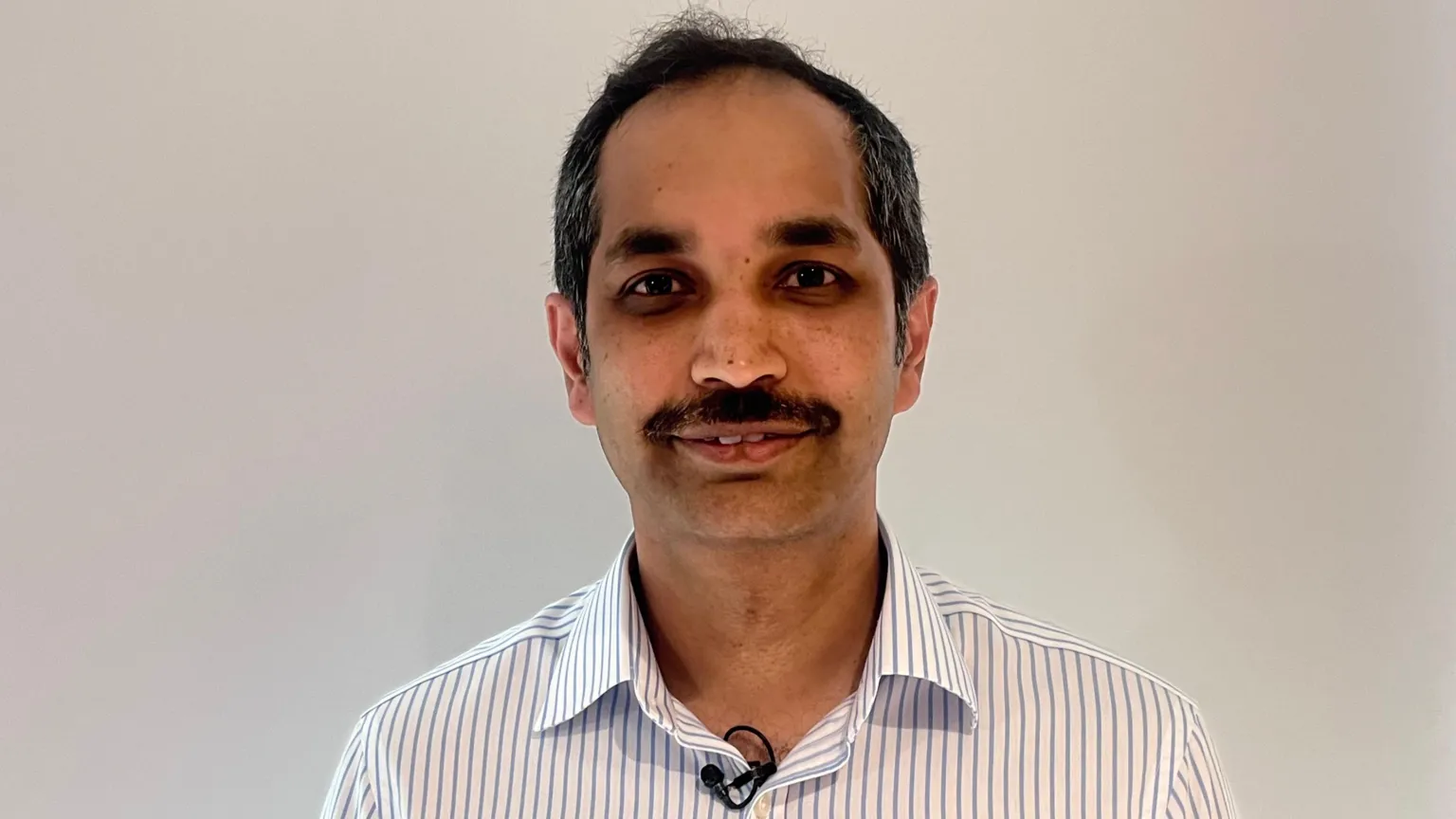 A man is standing in front of a white wall. He has tan skin and black hair. He also has a black moustache. He is wearing a white shirt with blue stripes. 