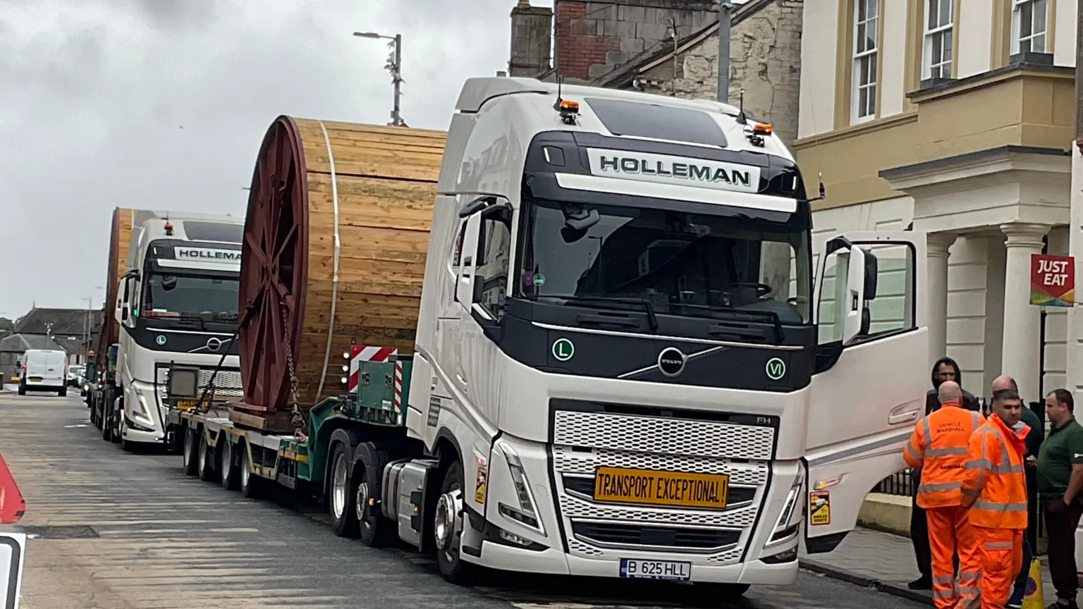 Charles Brown Two lorries in a row with large wooden wheels on the back of them. There are two men in orange work clothes talking to others who might be the drivers.