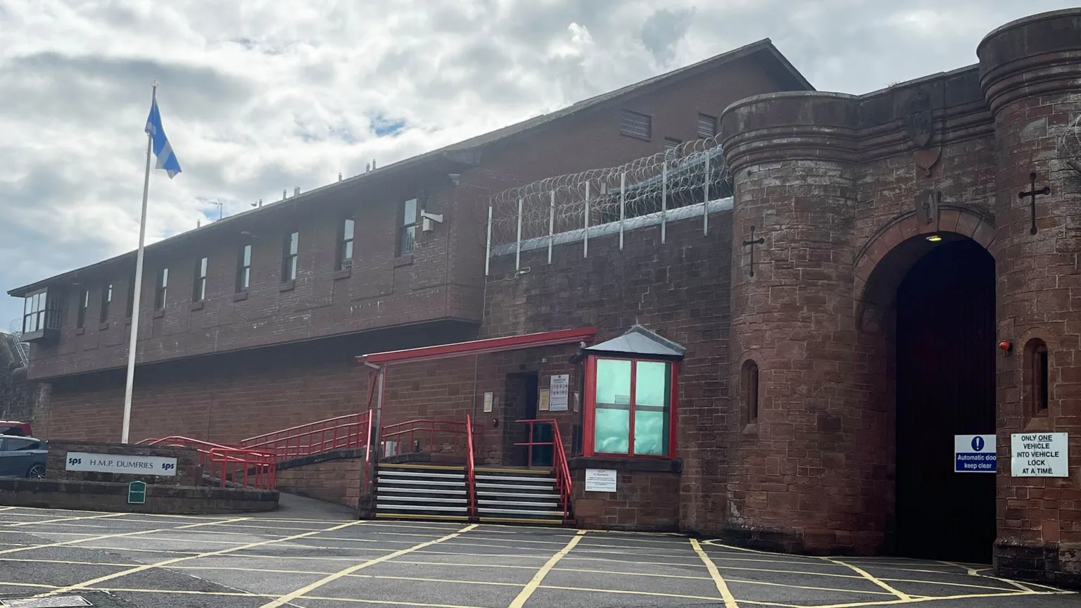 The exterior of Dumfries Prison, a red brick and sandstone building. There is a saltire flying on a flagpole to the left, next to steps with red handrails and a glass box at the entrance. The section on the right looks more like a castle entrance with a large arched opening.