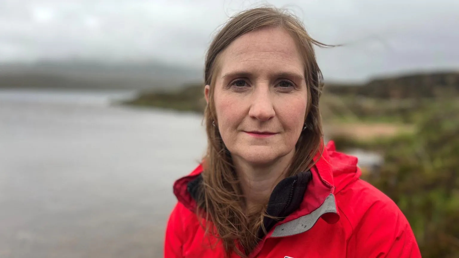 A woman with long brown hair and a red rain jacket. She is standing in front of a loch.