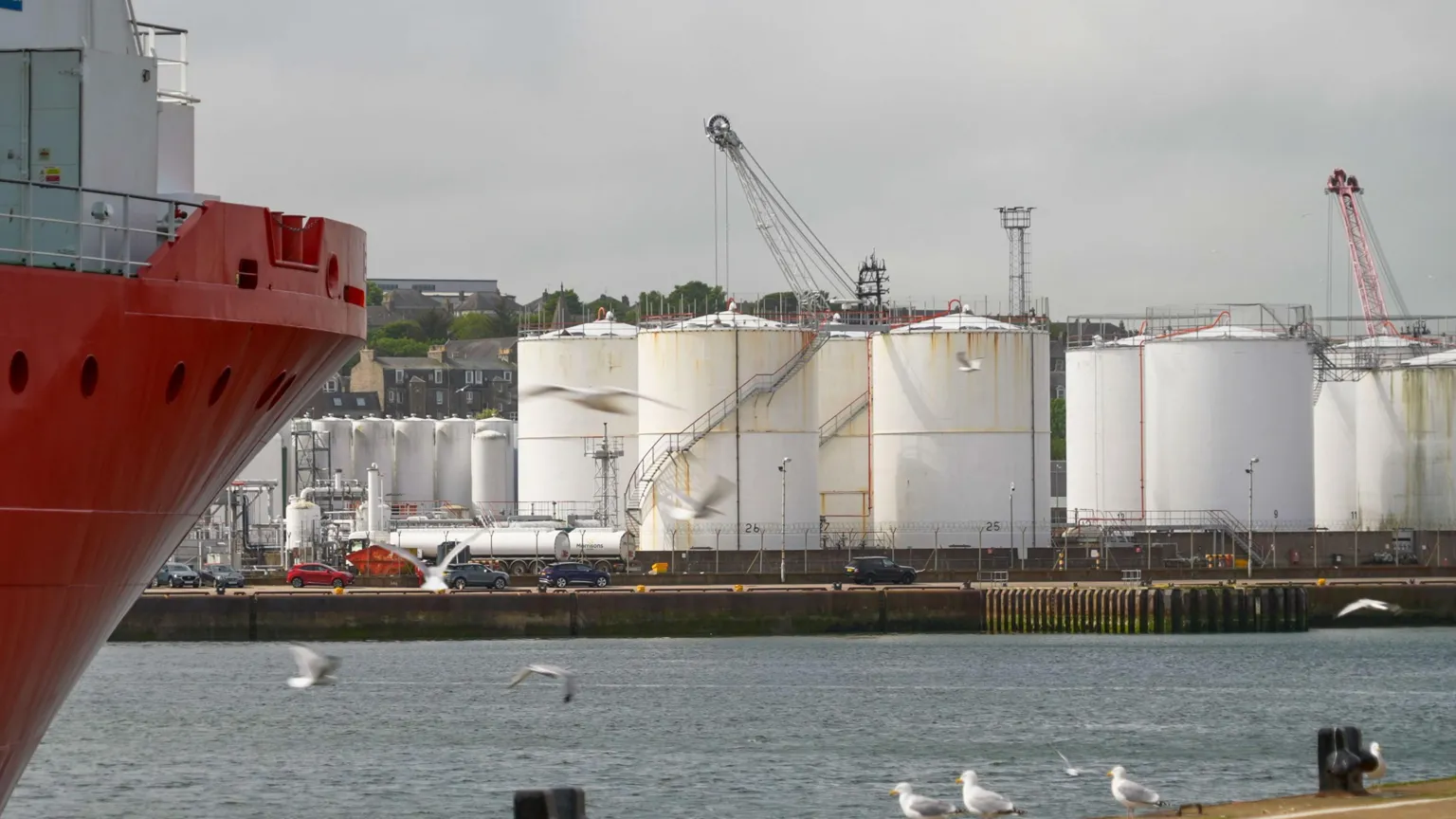 A harbour with a oil plant in the distance and seagulls flying in the foreground by the hull of a red boat