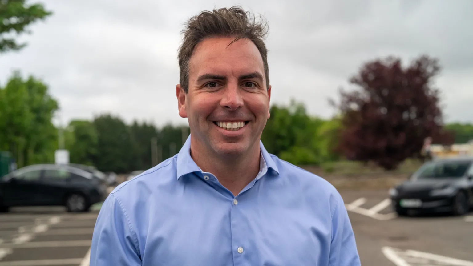 Maurice Golden, a dark-haired man in a light blue shirt, is in a car park and is smiling. The sky behind him is grey.