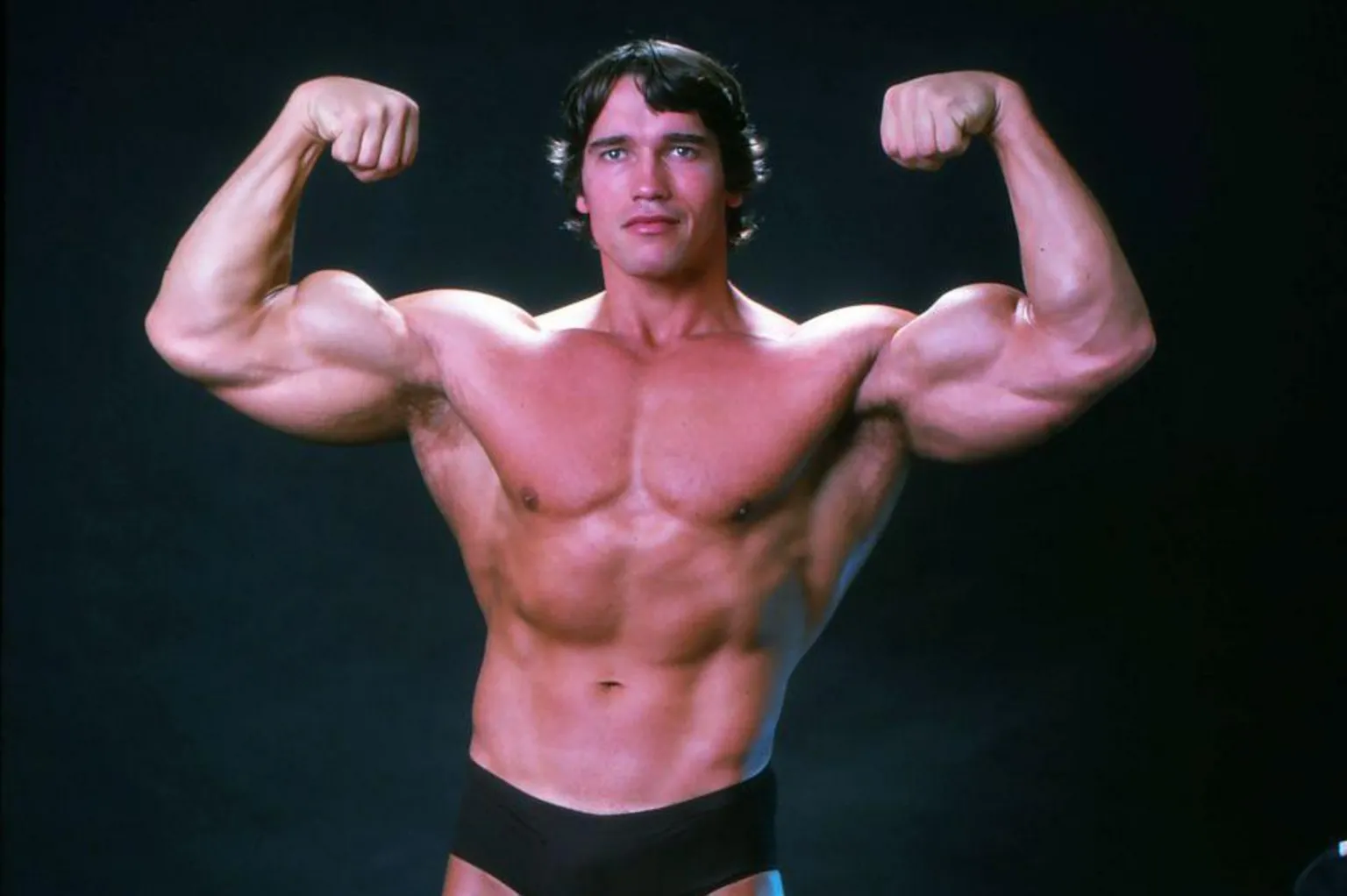  Arnold Schwarzenegger is his younger body-building days, he is wearing black trunks and is flexing his muscles.