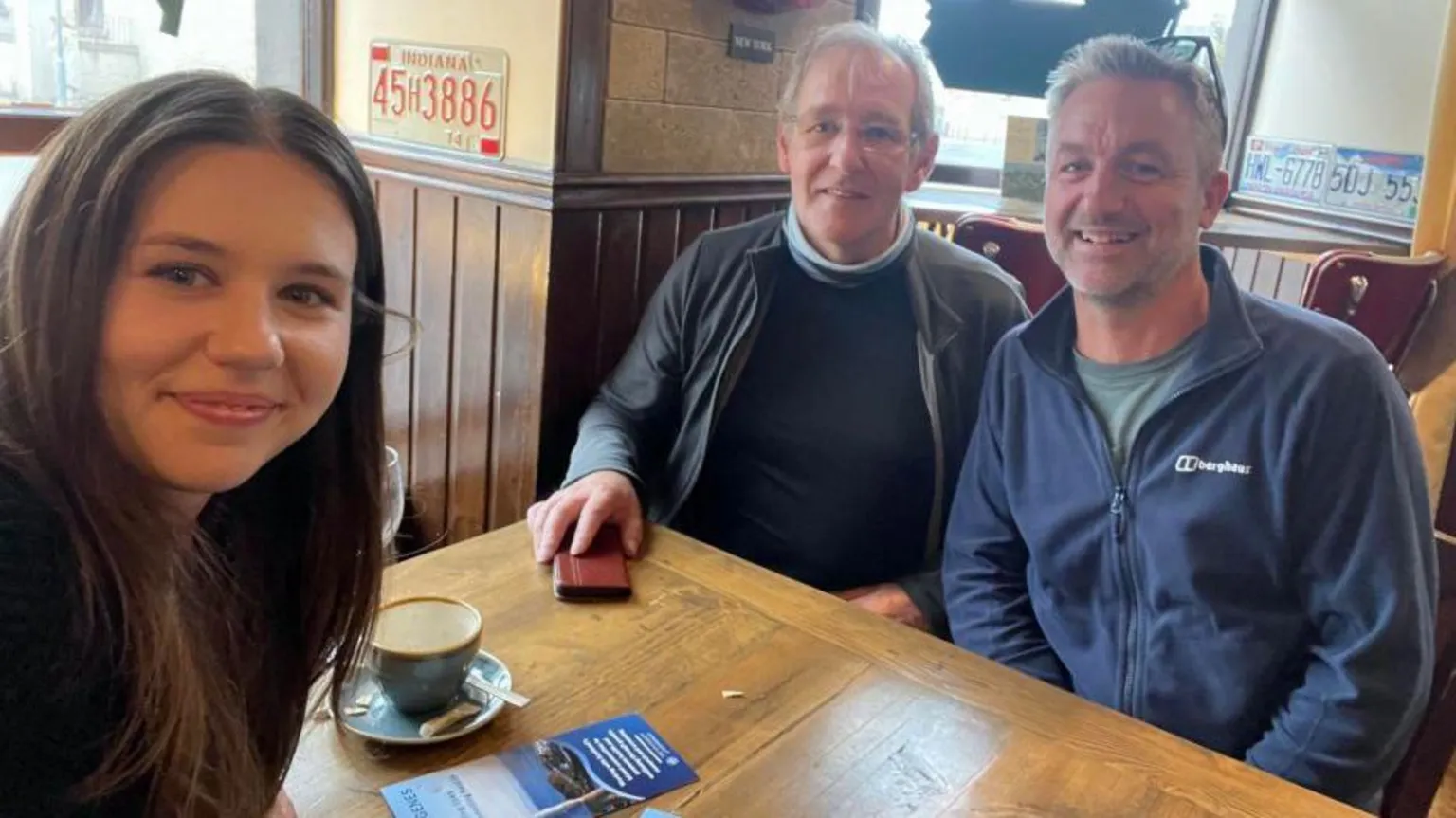 Flett family A young woman, Chloe Flett, smiling at the camera in a selfie in a cafe, with two men also sitting at the table - Stuart Flett, and Prof Jim Flett Wilson.