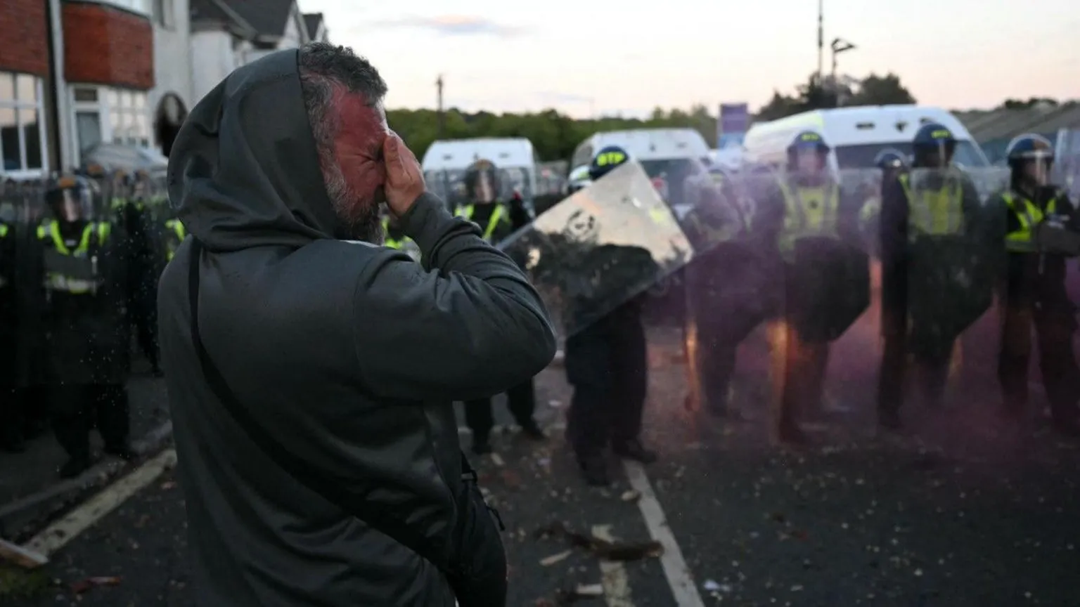  A man in a hooded jacket is pictured clasping his hand to his face. Behind him is a row of police officers holding riot shields. 