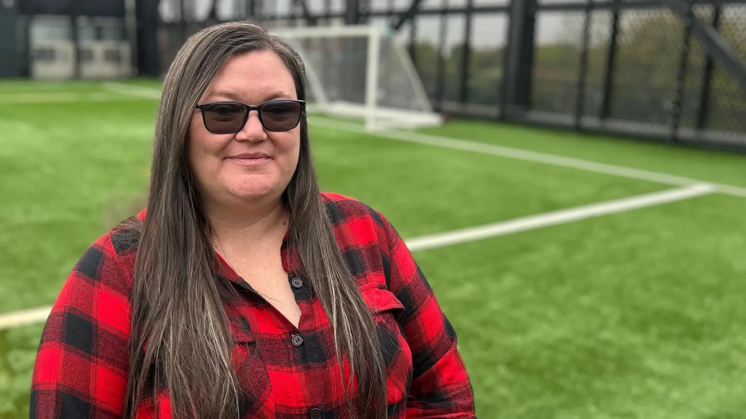 Sherrie Dalton is standing on an artificial football pitch with an out-of-focus goal behind her. She has long dark hair and is wearing prescription sunglasses and a red and black checked flannel shirt. She's smiling.