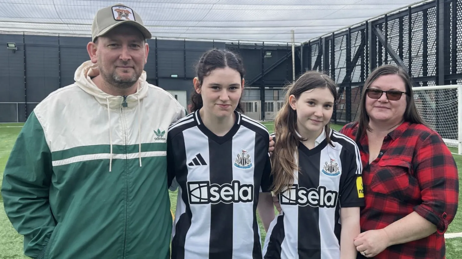David Dalton, Holl Dalton, Olivia Dalton and Sherrie Dalton stand on an astro turf football pitch. Mr Dalton is wearing a cream and green Adidas sports jacket and a Liverpool football cap. Holly and her sister are wearing Newcastle tops. Both girls have long dark hair. Sherrie Dalton is wearing a red and black checked flannel shirt. They are standing close together and smiling. 