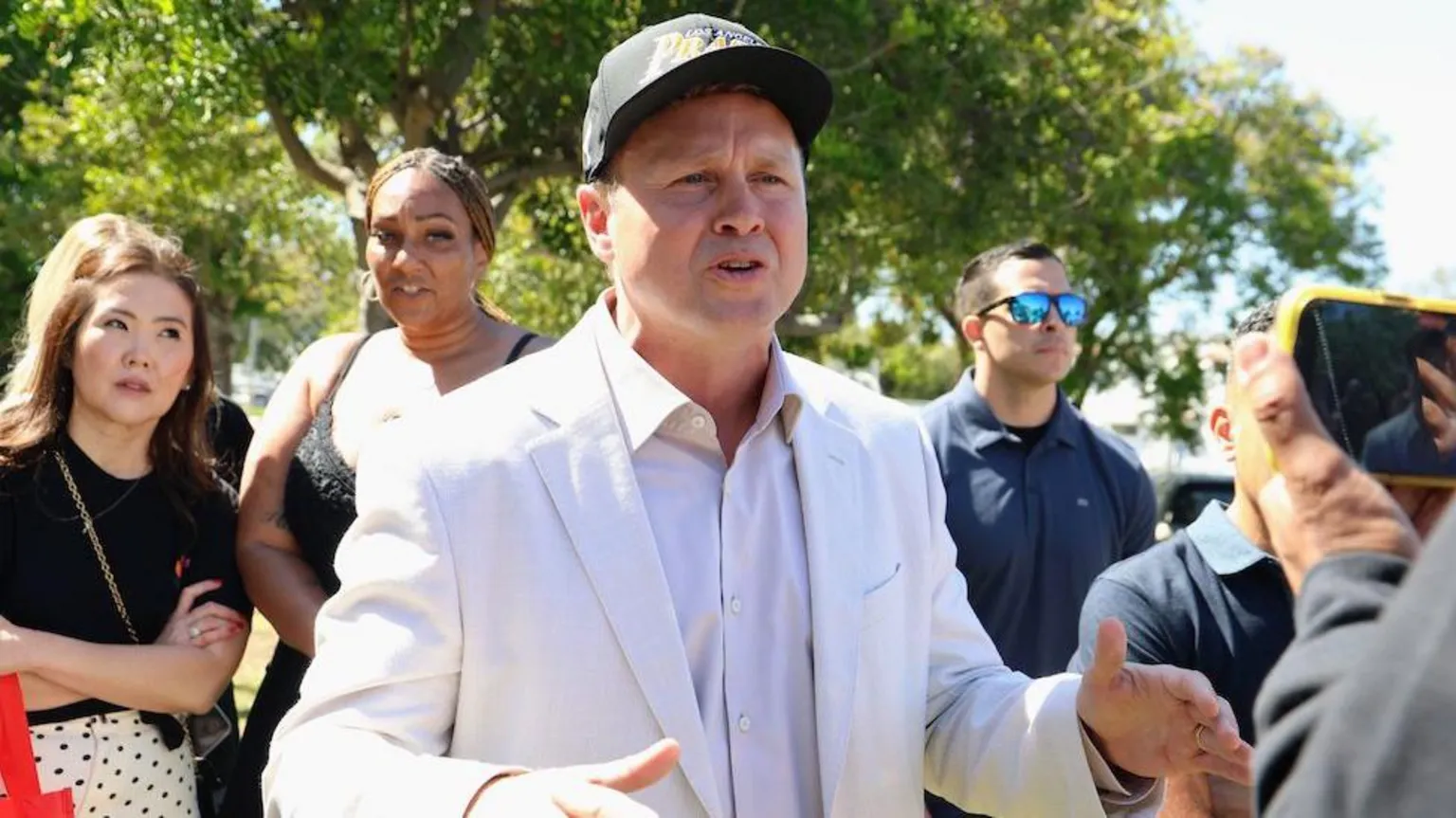  Spencer Pratt speaks to voters on election campaign. He is wearing a black baseball cap and a white linen suit and white shirt. There is a crowd gathered around him to hear him speak.