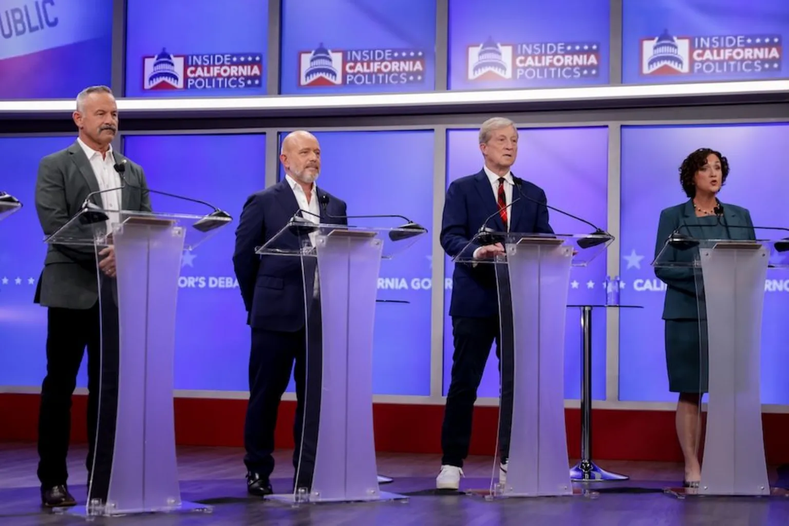  There are four people stood behind podiums on a stage. The two figures in the centre are among the leaders in the polls. Steve Hilton is bald and bearded and wearing a dark suit and white shirt. To his left is Tom Steyer who is wearing a dark suit, red tie and white trainers.