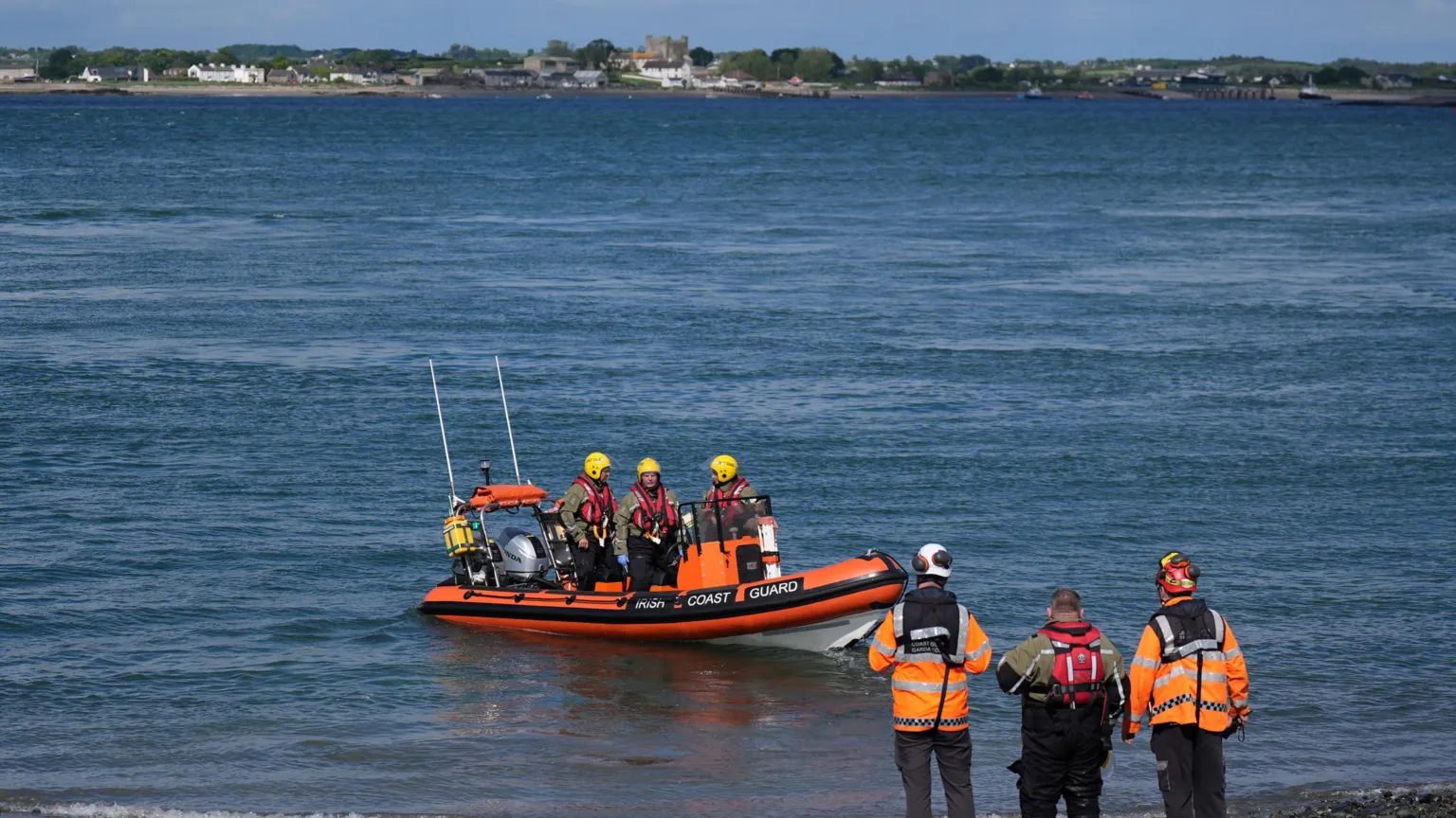  An Irish Coast Guard boat on a body of water, with some green fields in the background. There are three people on the orange inflatable boat, wearing red life jackets and yellow helmets. There are three people standing on the shore, waiting for the boat to come in. Two are in orange hi-vis jackets. One is wearing a red life vest.