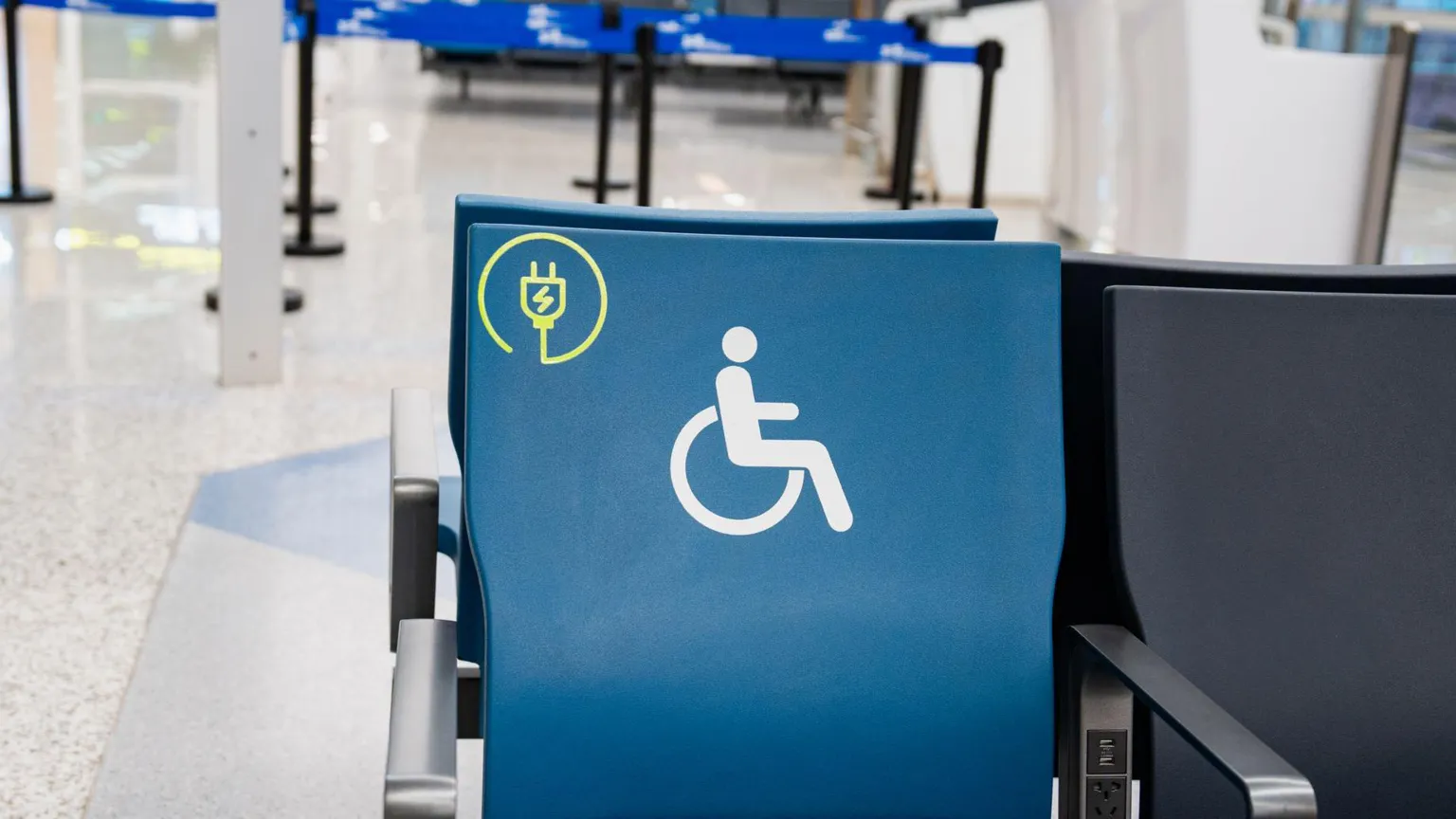  A blue chair with a disability sign on it it seen in an airport. Behind it, there are barriers leading up to a check-in desk.
