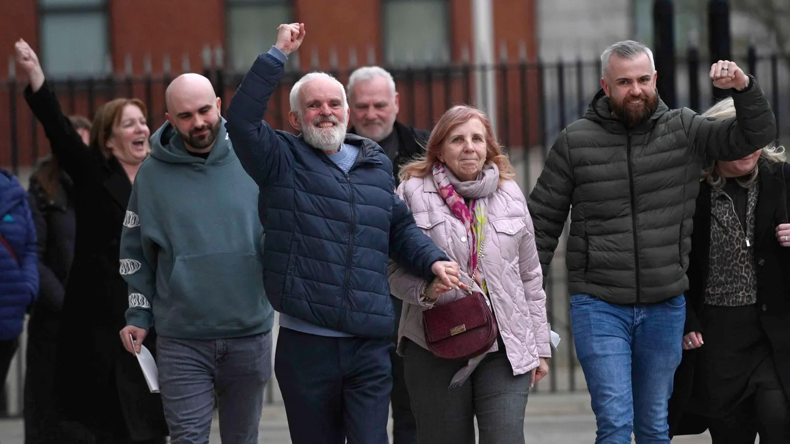  A group shot of seven people - three of whom have their arms raised in a celebratory way. The man in the centre - Natalie's dad - is wearing a blue puffer coat and has grey hair and a grey beard. He is holding hands with Natalie's mum who is wearing a pink/purple coat, with a matching scarf. 