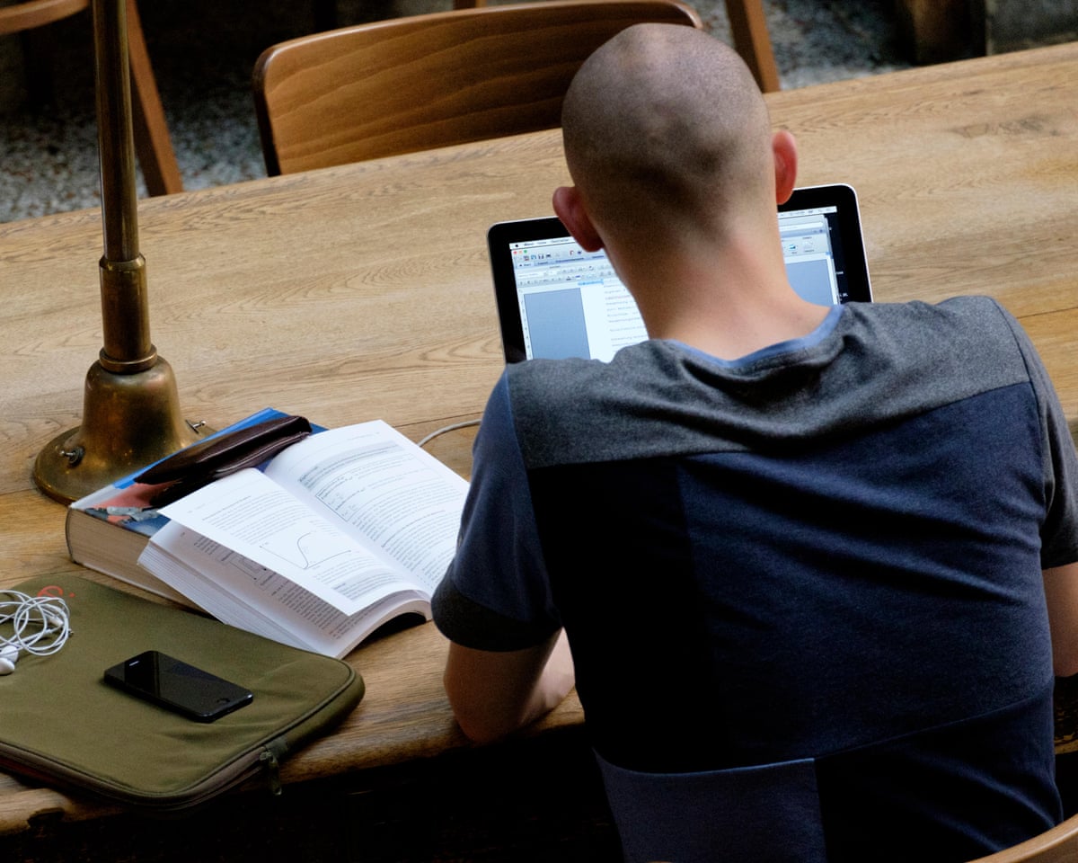 Student photographed from behind, sitting at a table on a laptop with an open book propper up next to them