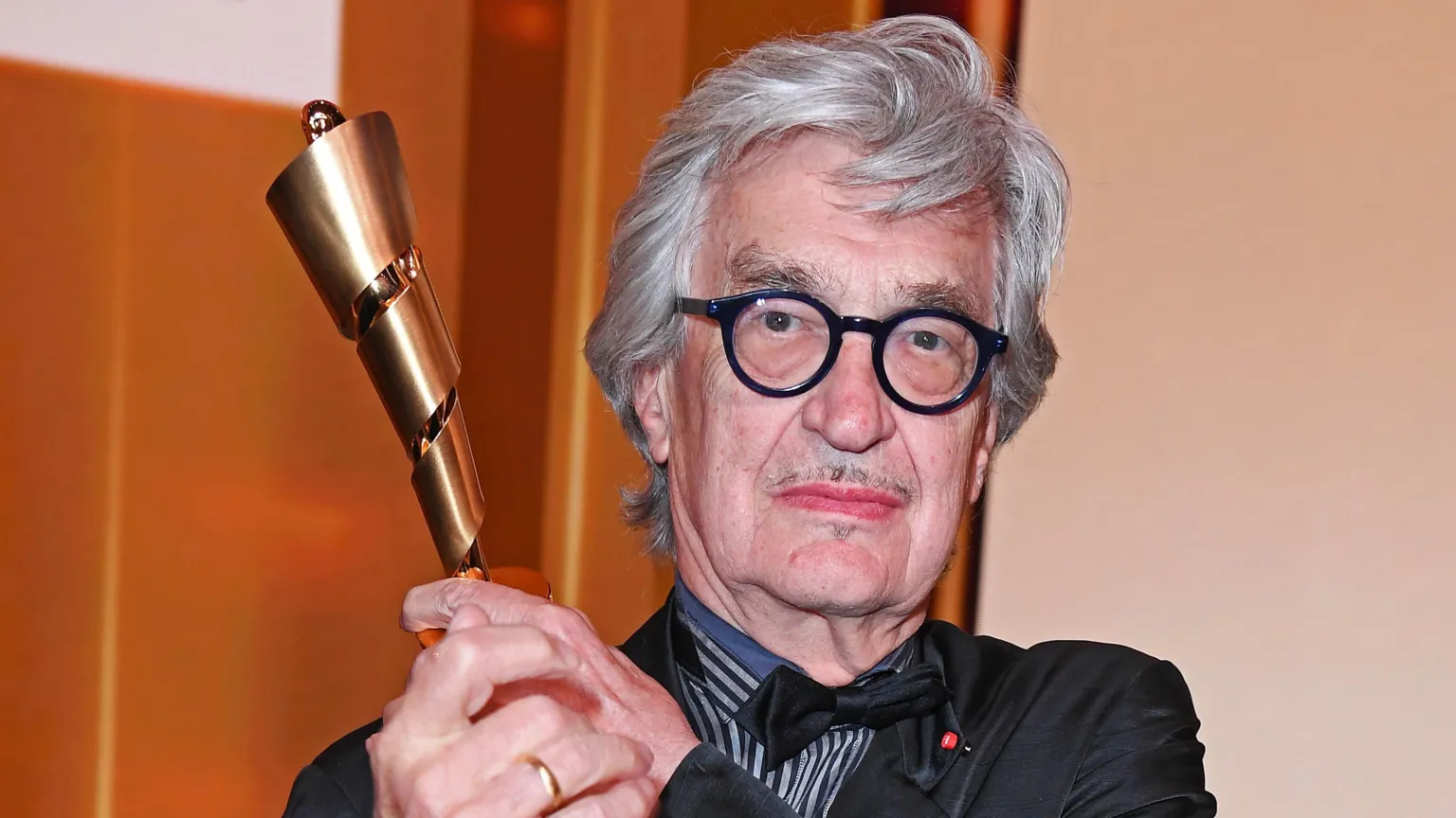 Wim Wenders with grey hair and black glasses holding up an award