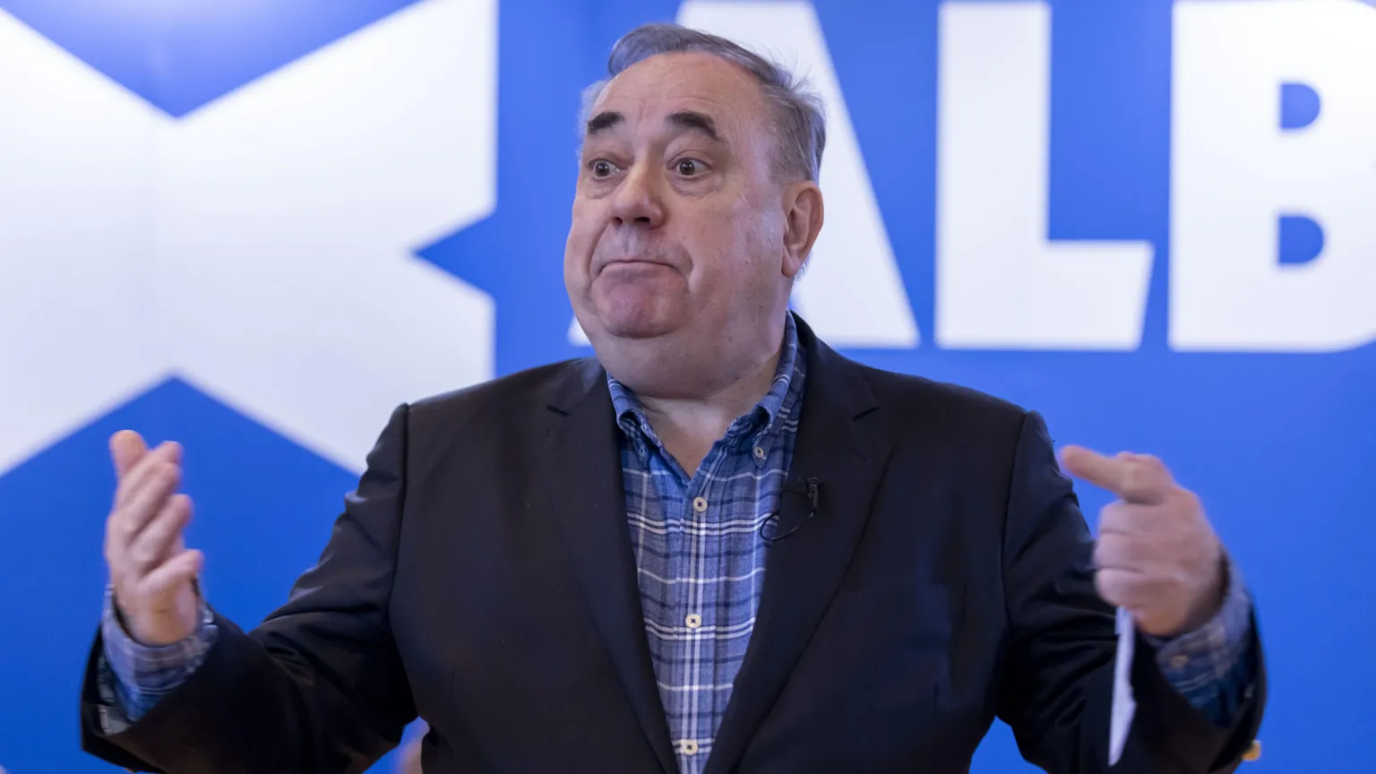 Alex Salmond standing in front of a blue background with the Alba Party logo on it.