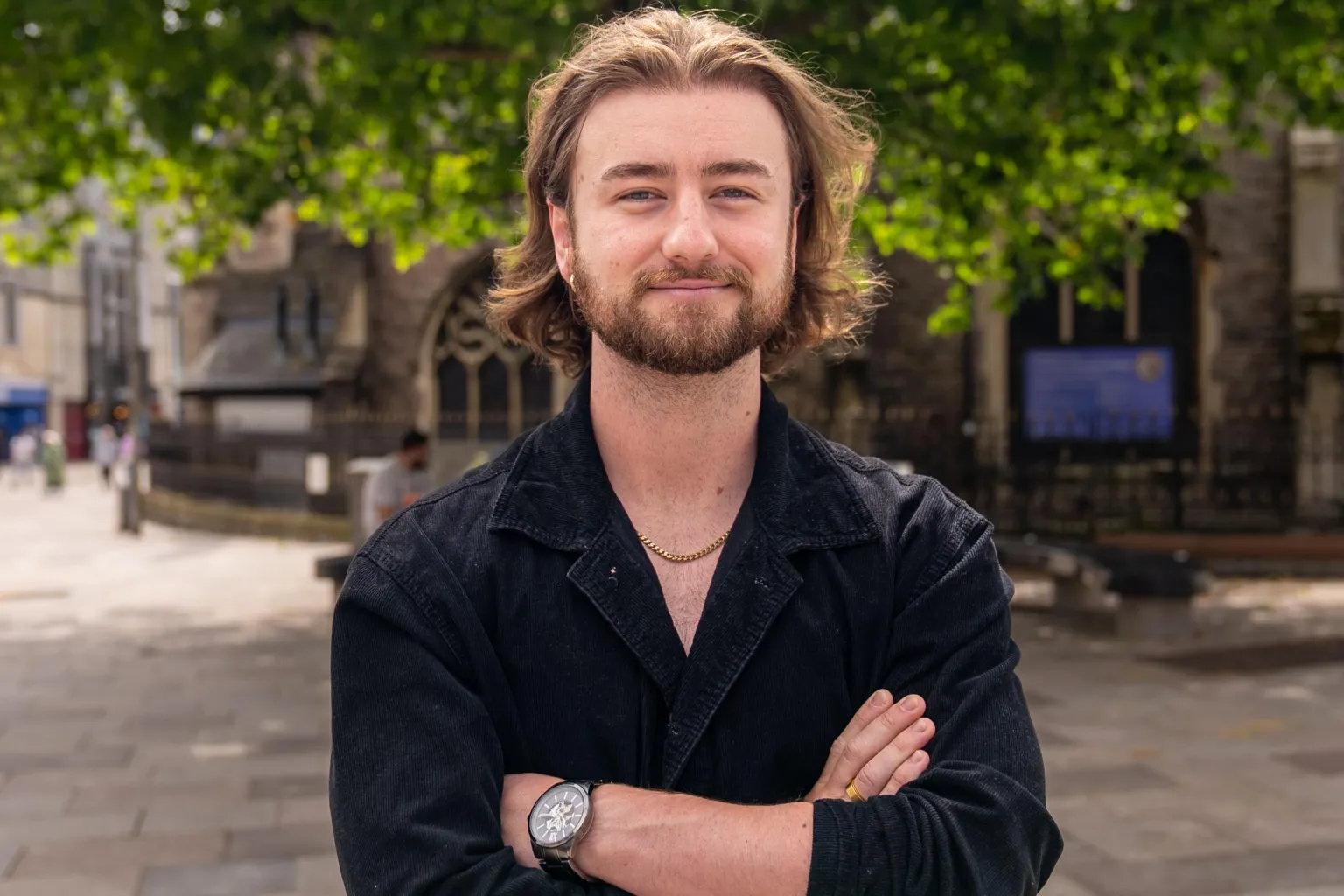 Tom crosses his arms stood in a city centre. He has a watch on his left wrist and a gold chain around his neck. He is wearing a black shirt and has long flowing brown hair.