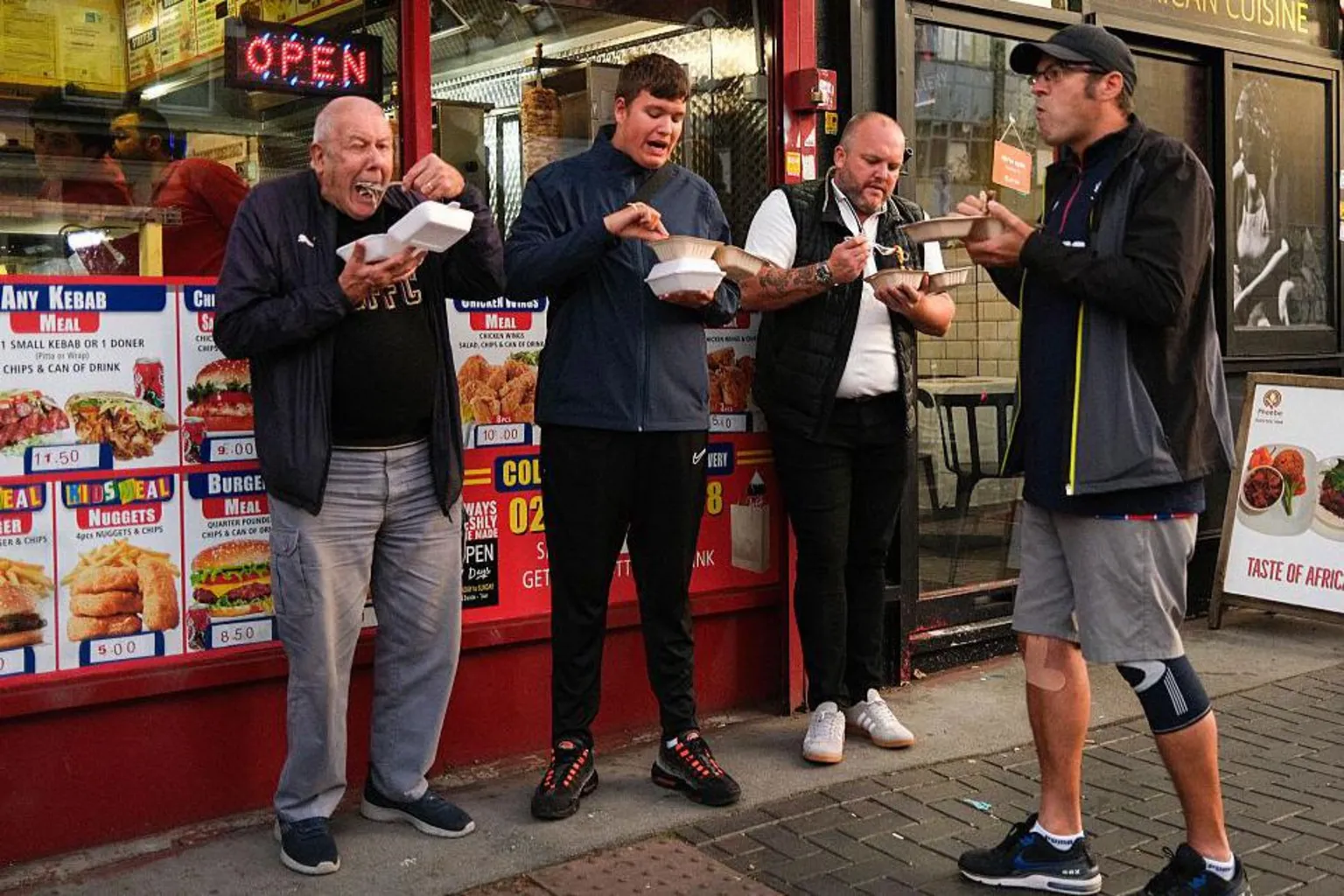 Four men stood outside a takeaway shop eating out of polystyrene white containers. There are images of food behind it