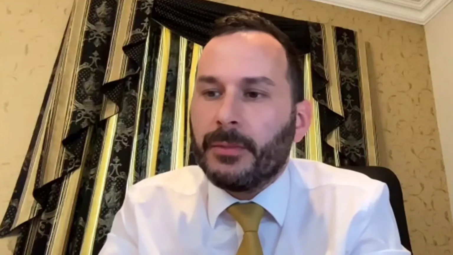 NI Assembly A man with black hair and beard wearing a white shirt and yellow tie sitting in a room with yellow wallpaper and in front of black curtain for a video call.