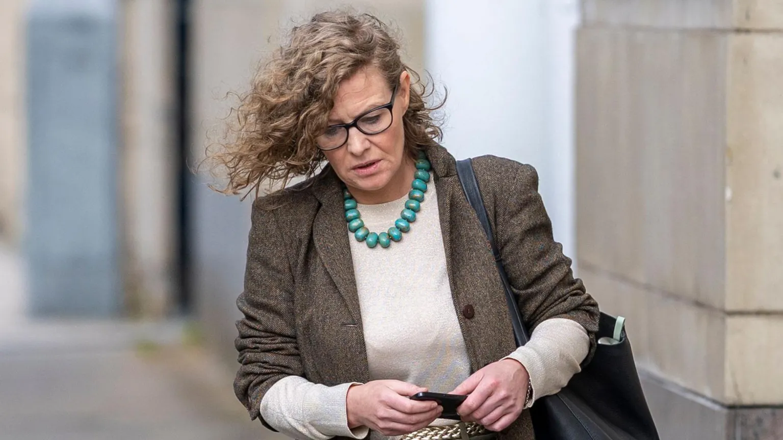 Liam McBurney/PA Wire Fiona Donohoe pictured looking towards the ground as she walked towards a court building in Belfast in April. She is a woman with shoulder-length, curly blonde hair. She is wearing black square-framed glasses, a brown tweed suit jacket with rolled-up sleeves, a crew-neck cream/beige top, a gold plaited belt, khaki trousers and a chunky turquoise beaded necklace. She is carrying a large black bag over one shoulder and is holding a black mobile phone in both hands.