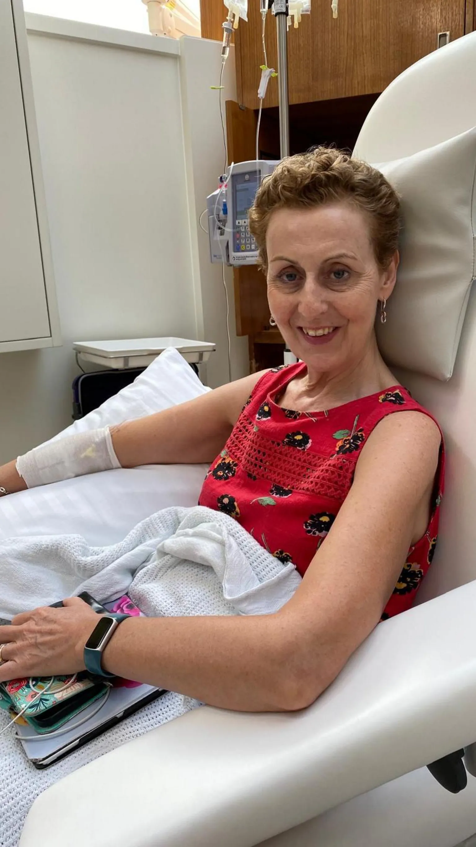 Patricia Hill The image shows a person sitting in a reclining medical chair in a clinical setting. The person is wearing a sleeveless red top with a patterned design and has a blanket draped over their lap. An intravenous line is inserted into the person’s arm, secured with a bandage, and connected to medical equipment mounted behind the chair. The person’s arm rests on the chair’s armrest, and a small bag or personal items are placed nearby. The room has clean white and wooden surfaces, with medical devices and monitors visible in the background. 