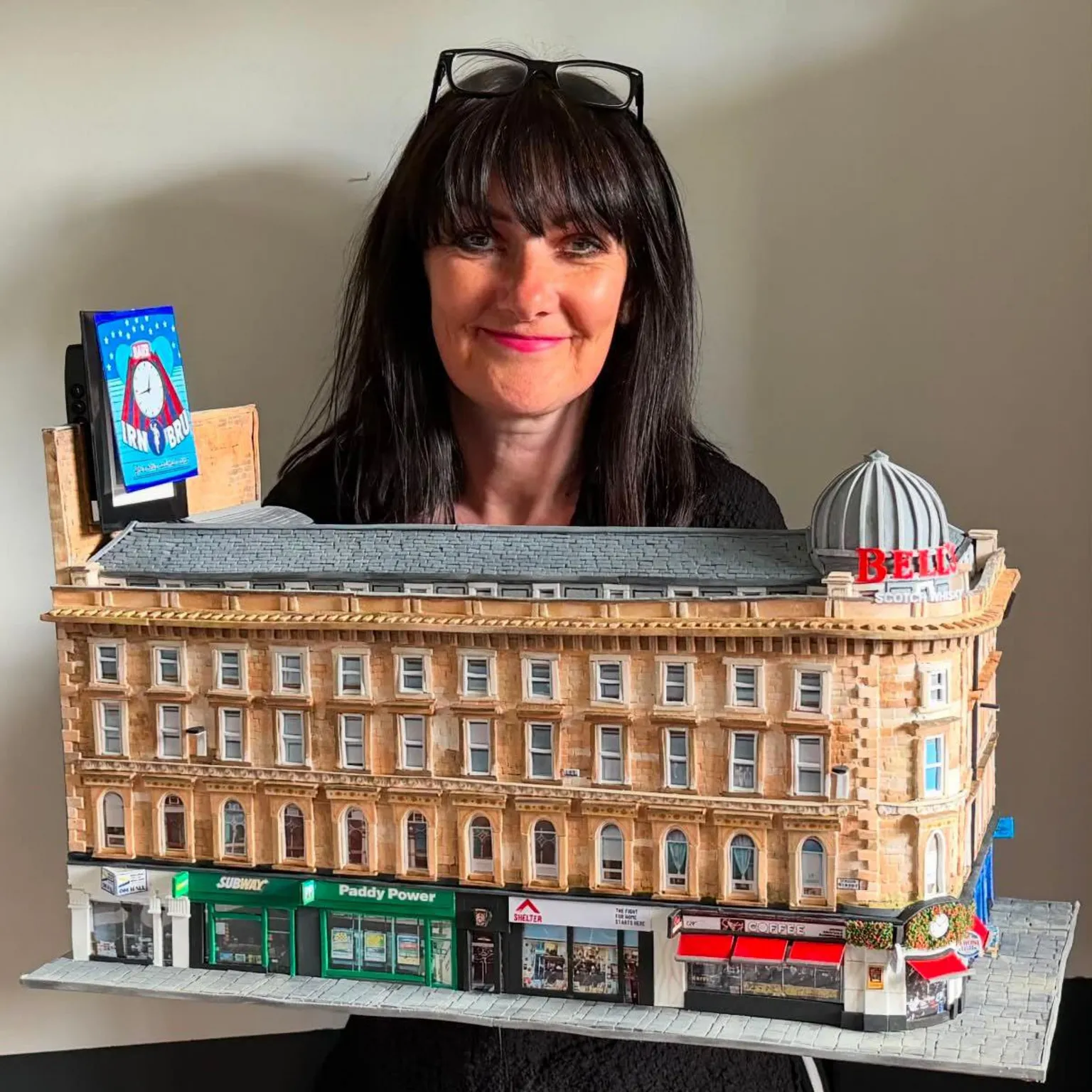 Karen Bones Karen, a woman in her 50s with dark brown hair and glasses on her head, holding the model of Union Corner.