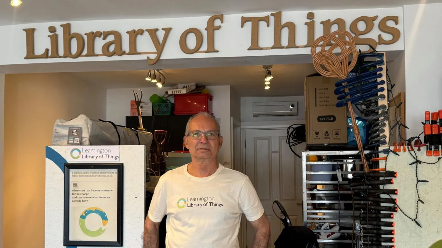 A man in a white t-shirt with Leamington Library of Things written on the front stands in front of a counter in a shop with the words library of things above the counter.