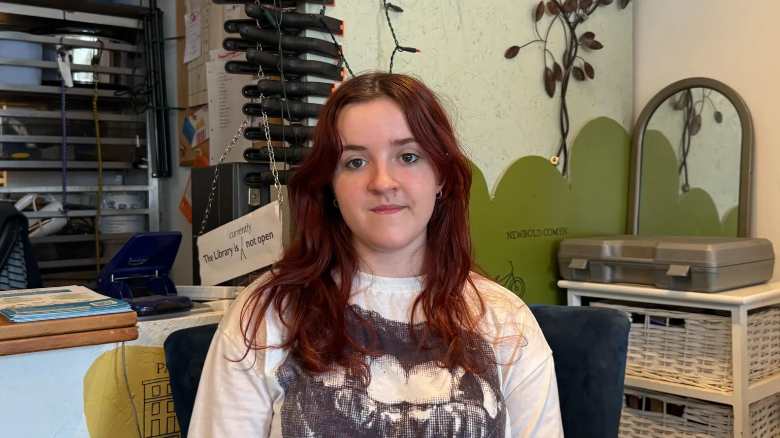 A young woman with long dyed red hair and brown eyes sits on a dark blue chair with white basket draws behind her and fairy lights on the wall.