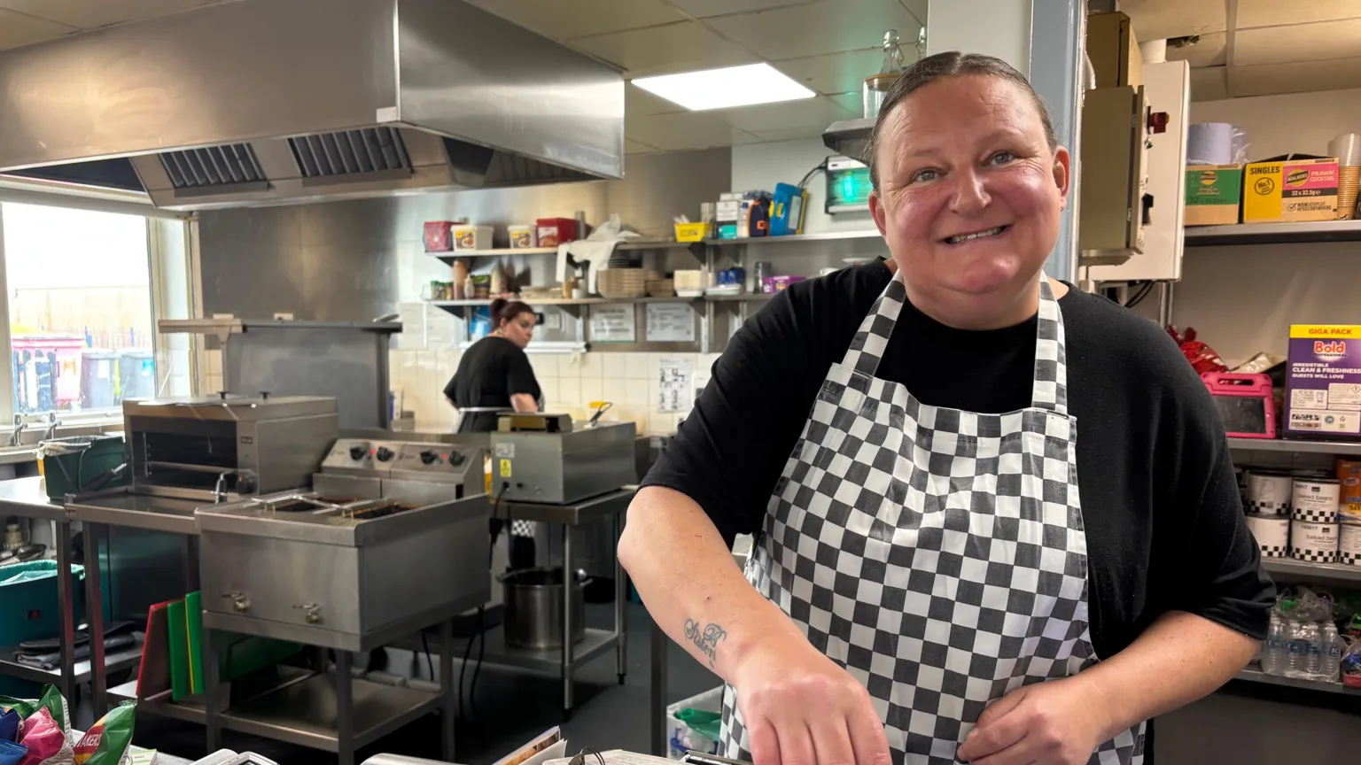 Leanna Russell who is wearing a checked chef's apron smiles as she stands in a large kitchen. Behind her are ovens and a fryer