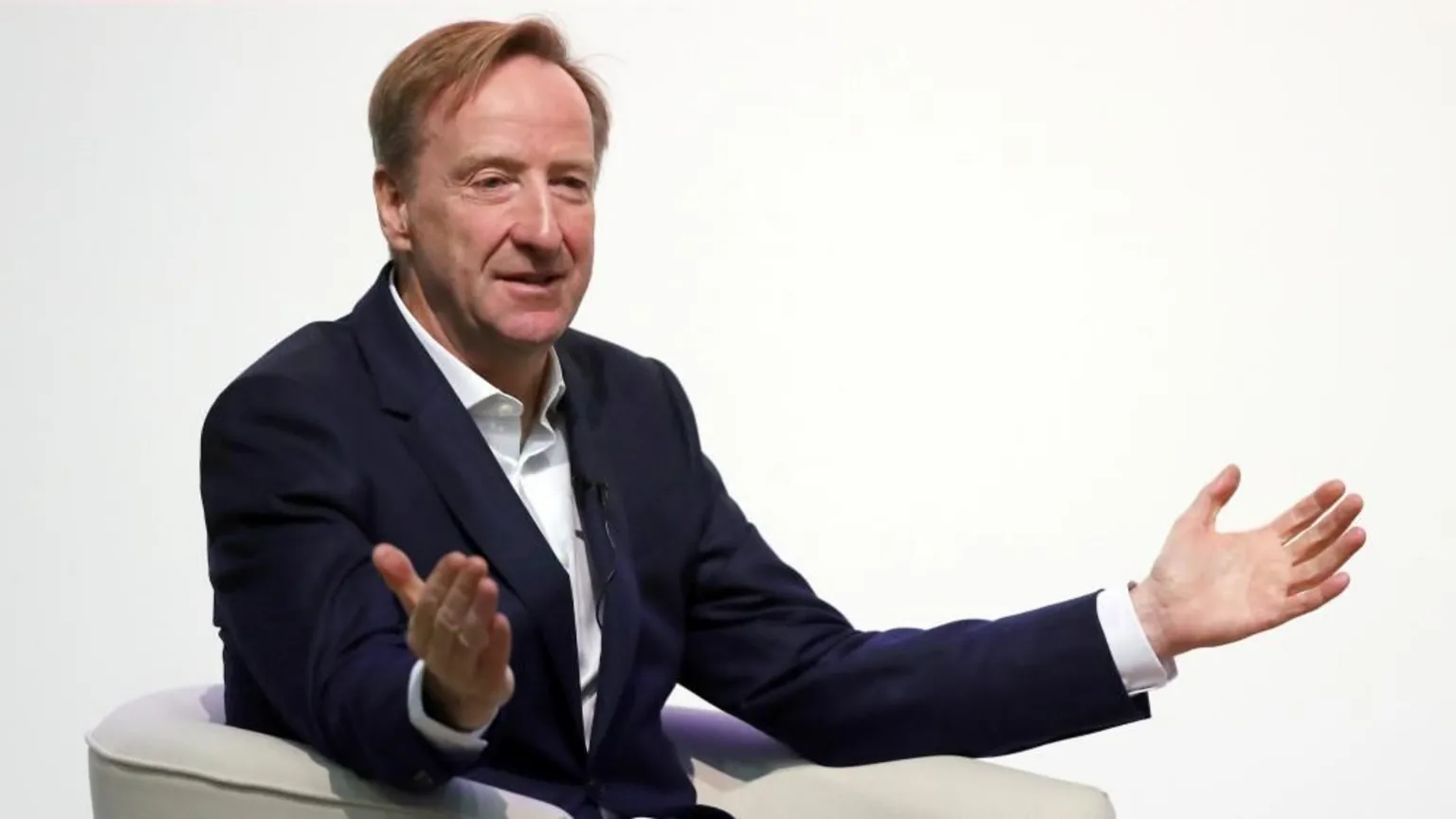  An older man with light, brown hair, dressed in a dark suit and white shirt and gesturing with open arms while sitting in a white chair.