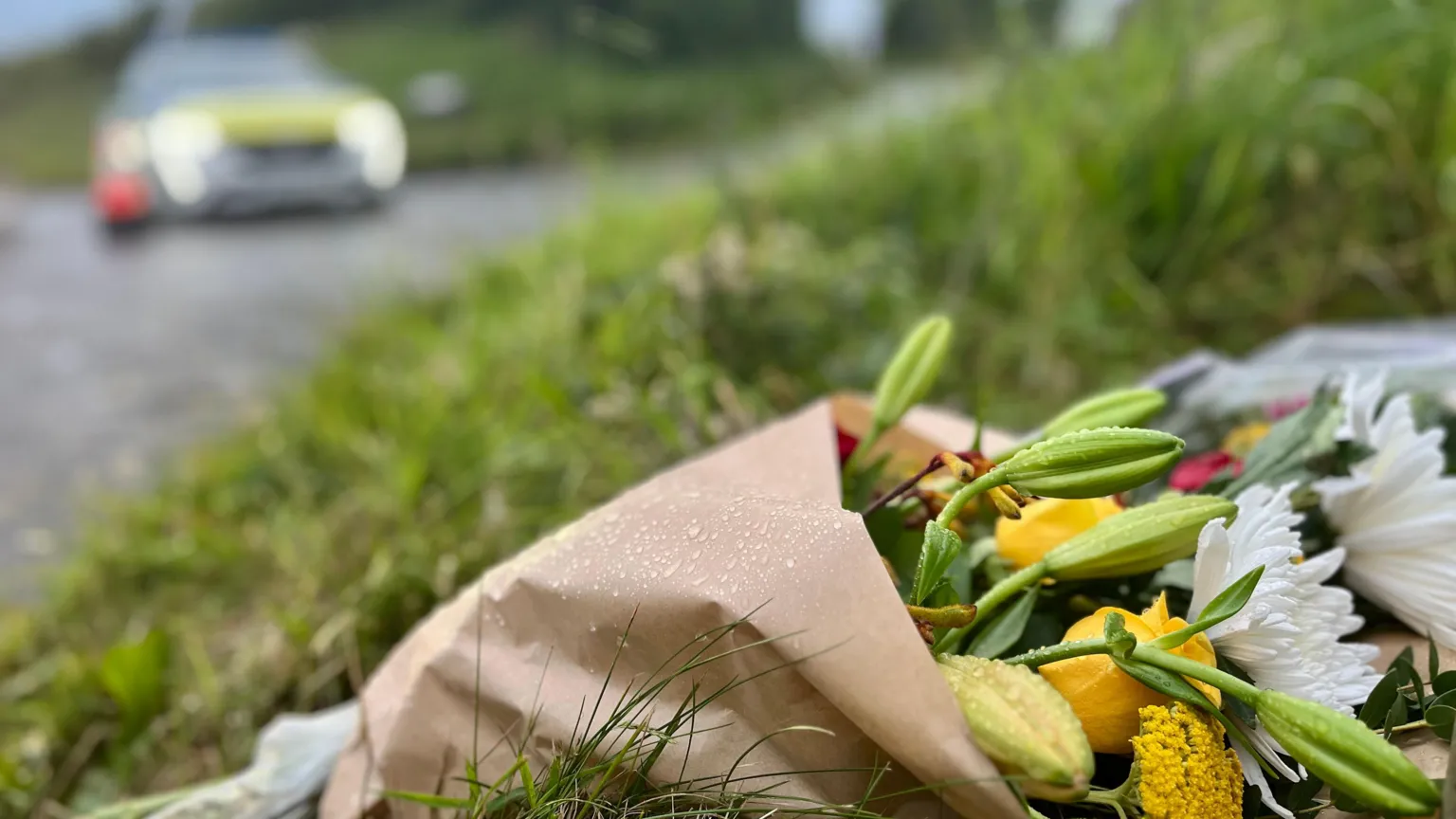Close-up shot of a bouquet of flowers left at the scene of a helicopter crash in Devon. A police car is blurred in the background.