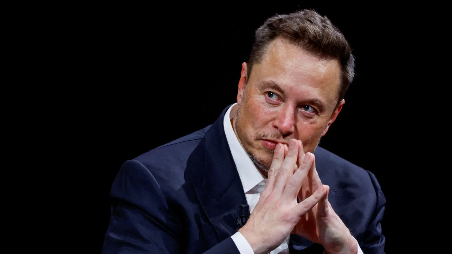 Elon Musk sitting down wearing a white shirt and navy blazer. He is looking to the left of the frame and has his hands clasped together in front of his mouth. He has short dark hair and stubble around his face.