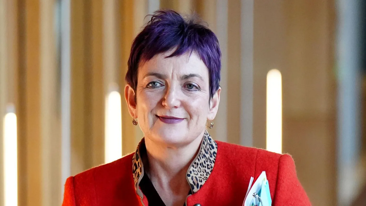  Angela Constance has short purple hair and wears a red blazer with a leopard print collar. She smiles and walks down a corridor at the Scottish Parliament