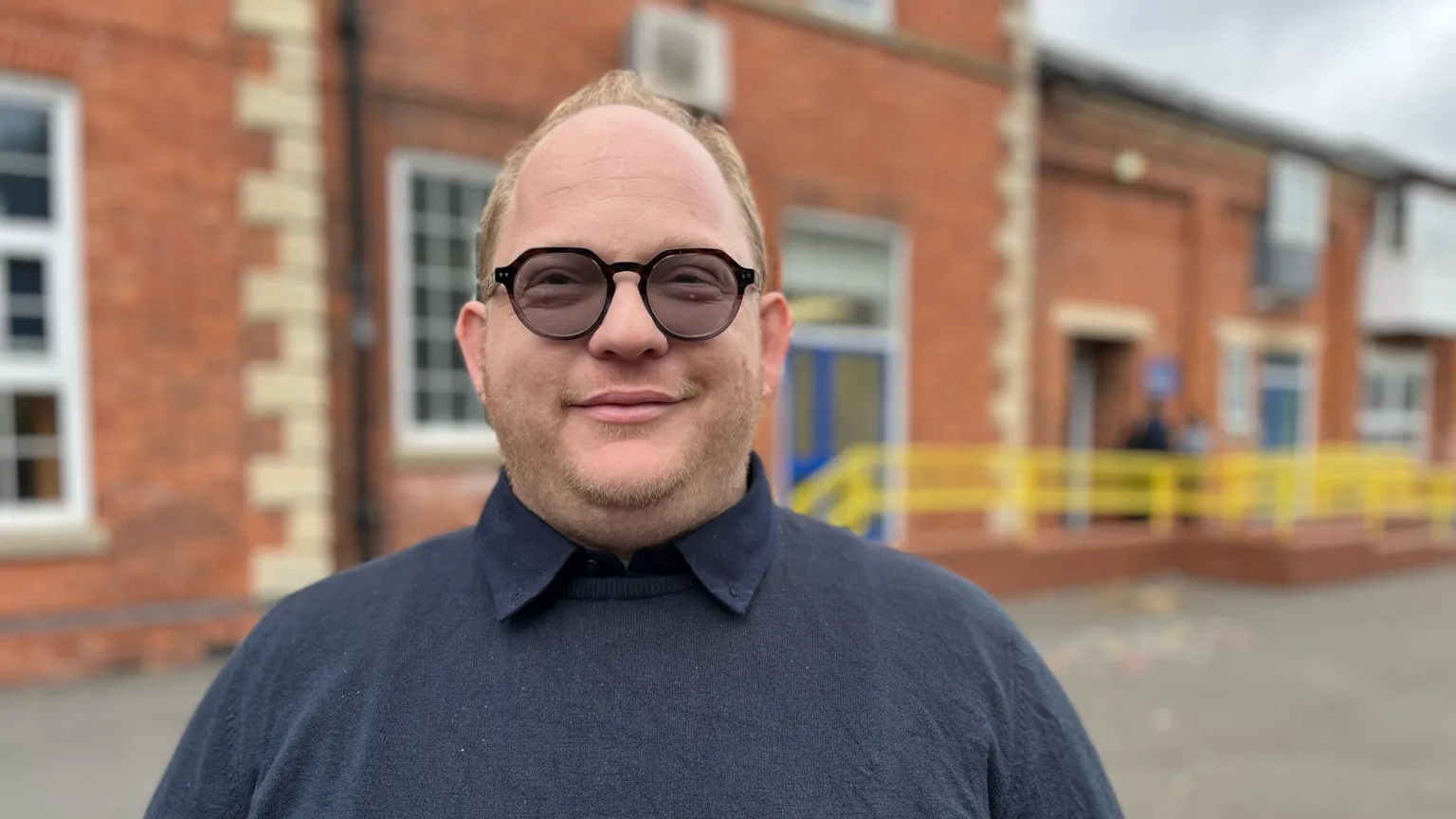 A man wearing a navy jumper and black glasses smiling, he has light blonde hair and a beard. 