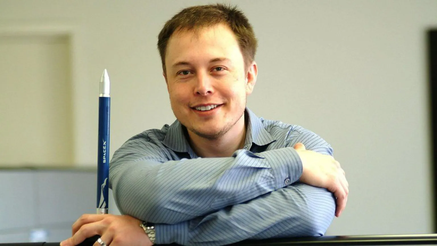  Elon Musk shown in 2004 wearing a blue shirt and smiling as he looks at the camera. He is holding a small, blue and silver toy rocket which bears the 
