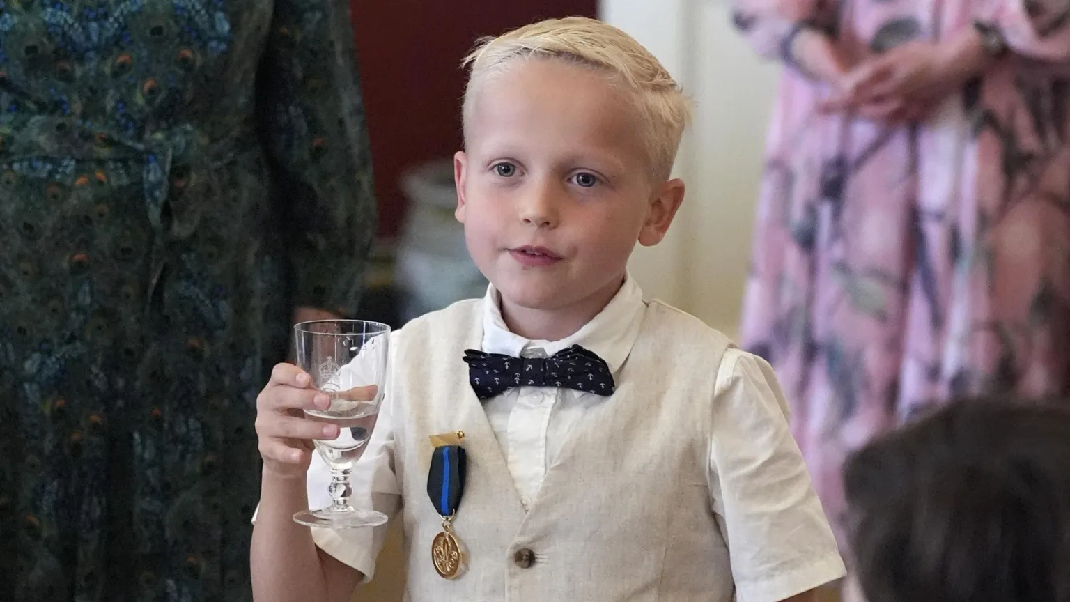  Rupert Williams with a glass of water. He wears a white short sleeved shirt, a beige waistcoat and a black bow tie. He has short blond hair. 