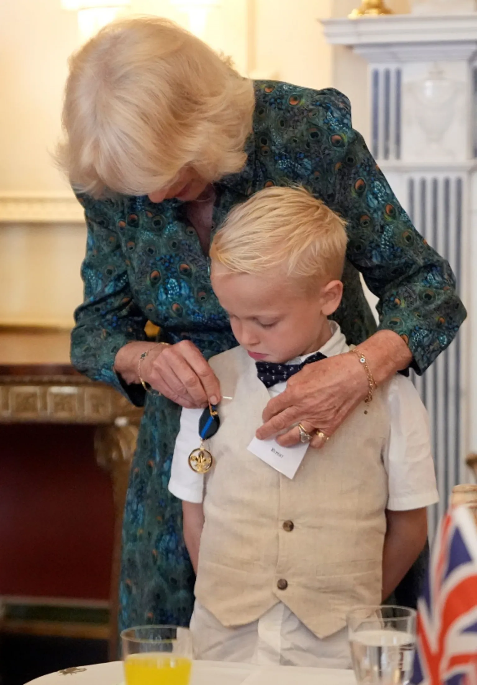  Rupert Williams standing in front of Queen Camilla. Rupert wears a white short sleeved shirt, a beige waistcoat and a black bow tie. Queen Camilla wears a patterned green, blue and orange dress. The Queen is pinning the medal on him