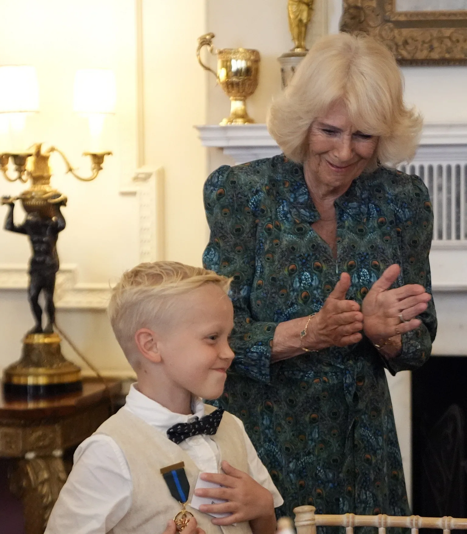  Rupert Williams standing in front of Queen Camilla. Rupert wears a white short sleeved shirt, a beige waistcoat and a black bow tie. Queen Camilla wearing a patterned green, blue and orange dress. Rupert smiles as his medal is pinned to his shirt. Camilla appears to be clapping. 