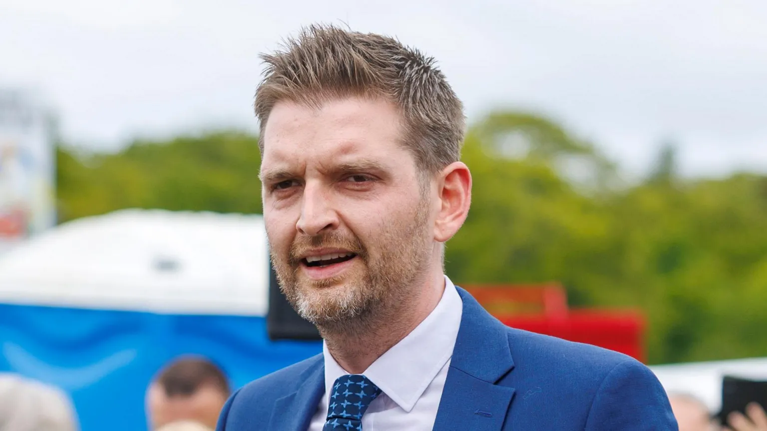  A man in a blue suit white shirt and tie standing outside. There is a gathering behind him which is slightly out of frame and focus.