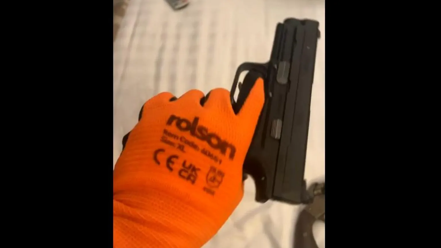 Counter Terrorism Policing An orange-gloved hand holding a black pistol. An old revolver is lying on the bed behind. 