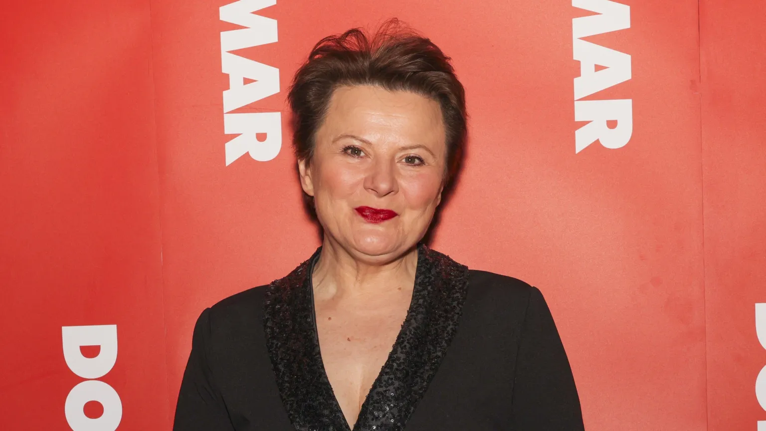 Monica Dolan wearing a black top and smiles at the camera. She is standing in front of a red background.