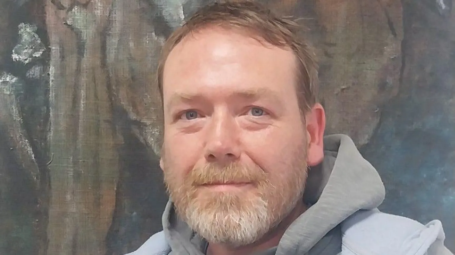 Irish Peatland Conservation Council Tristram Whyte, a man with blue eyes, short blonde hair and a greying beard, looks to the camera. He is wearing a grey hoodie under a grey zip jacket with a light blue fleeced lining. He is standing in front of a painting which is only partially visible