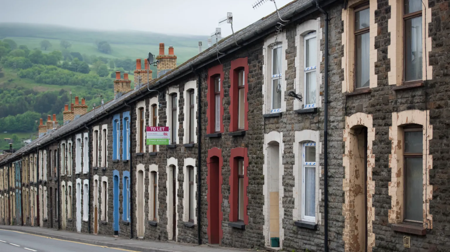  A row of terraced homes on a sloped street. They all have stone facades, but have different coloured window sills and porches. 