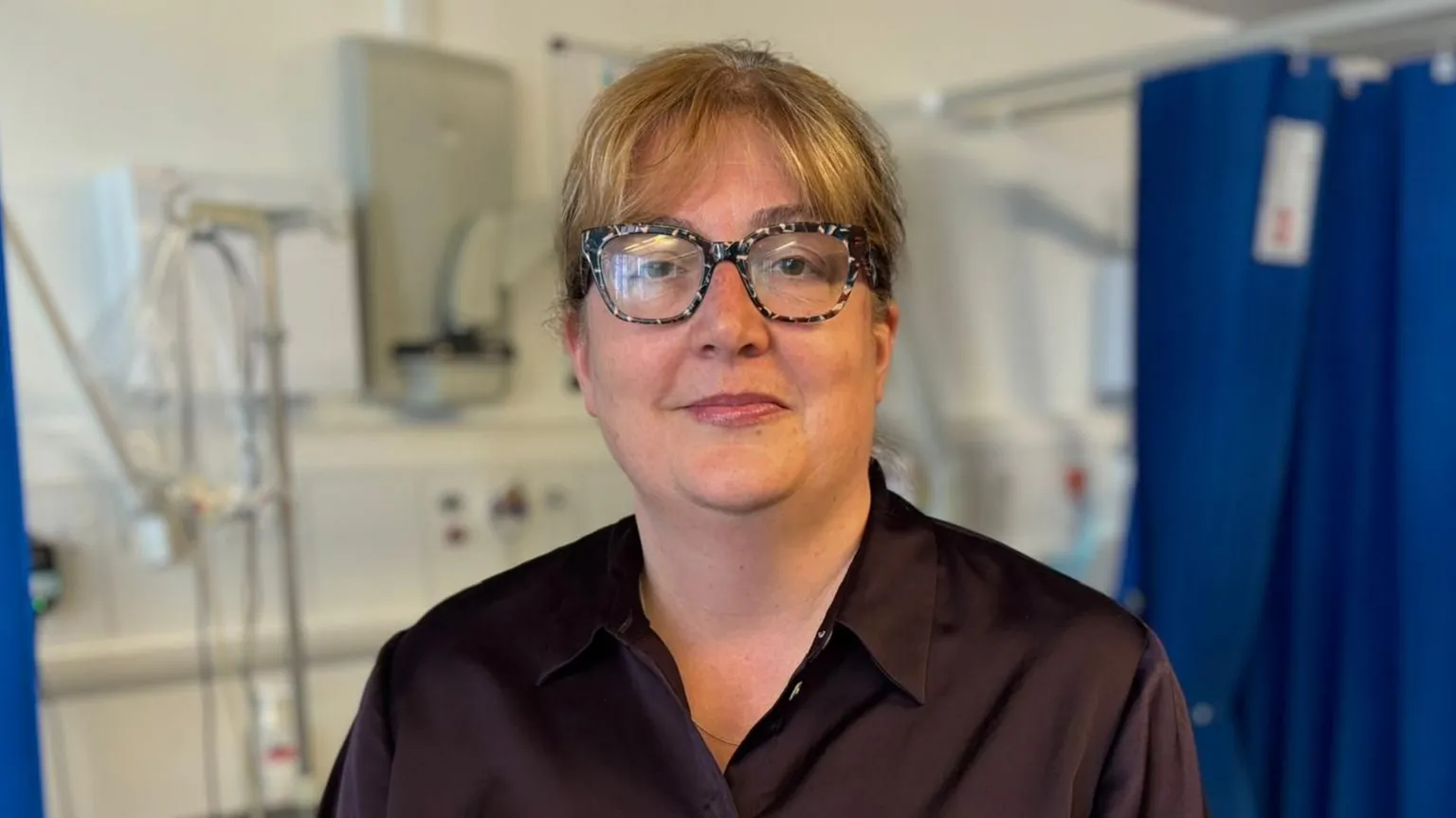 Portrait of a Justine wearing patterned glasses and a dark shirt, standing in a clinical room with medical equipment and blue privacy curtains visible in the background.