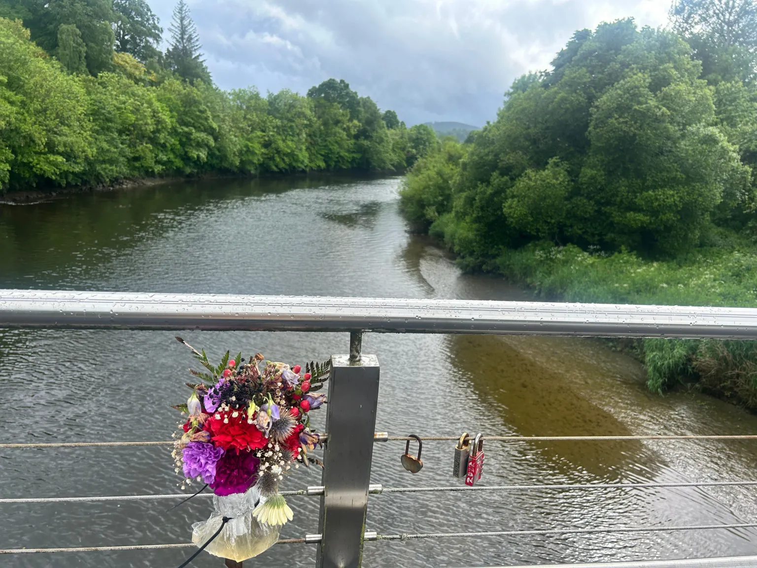 A bunch of flowers attached to a bridge over a river. There are full, green trees growing on the banks of the river. 