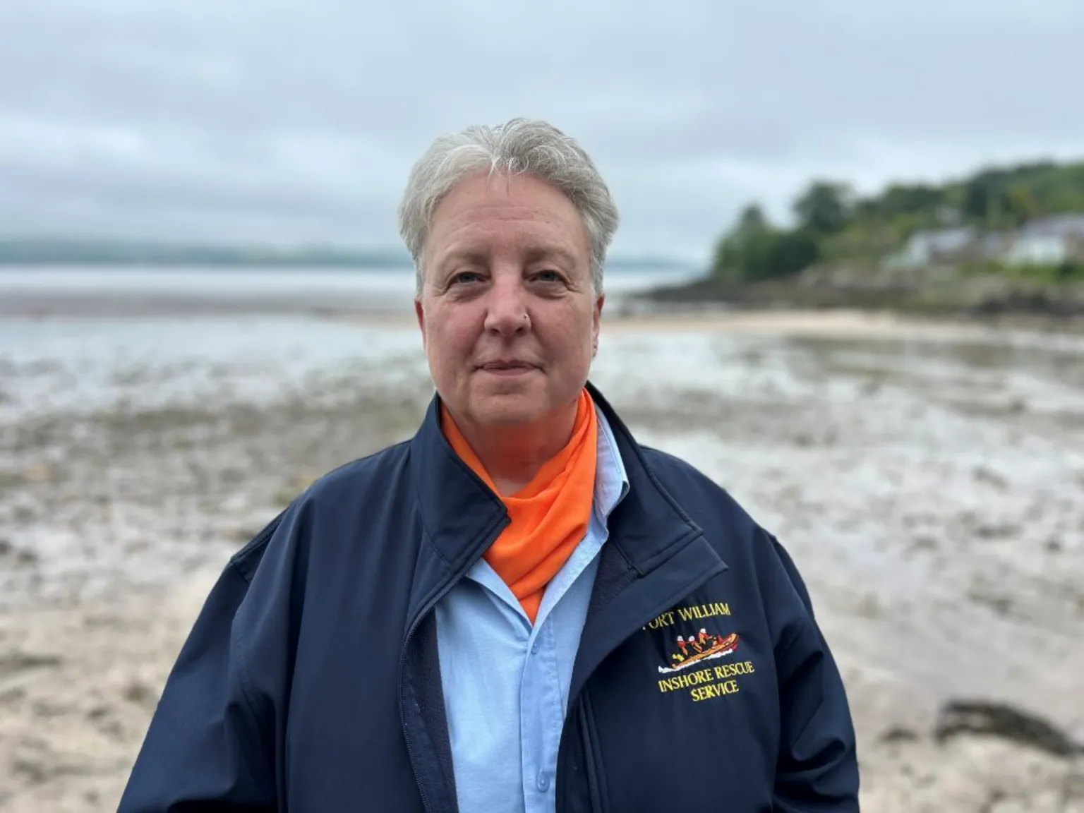 A woman with short grey hair standing on a sandy/muddy beach. She is wearing a blue jacket with a motif which says 