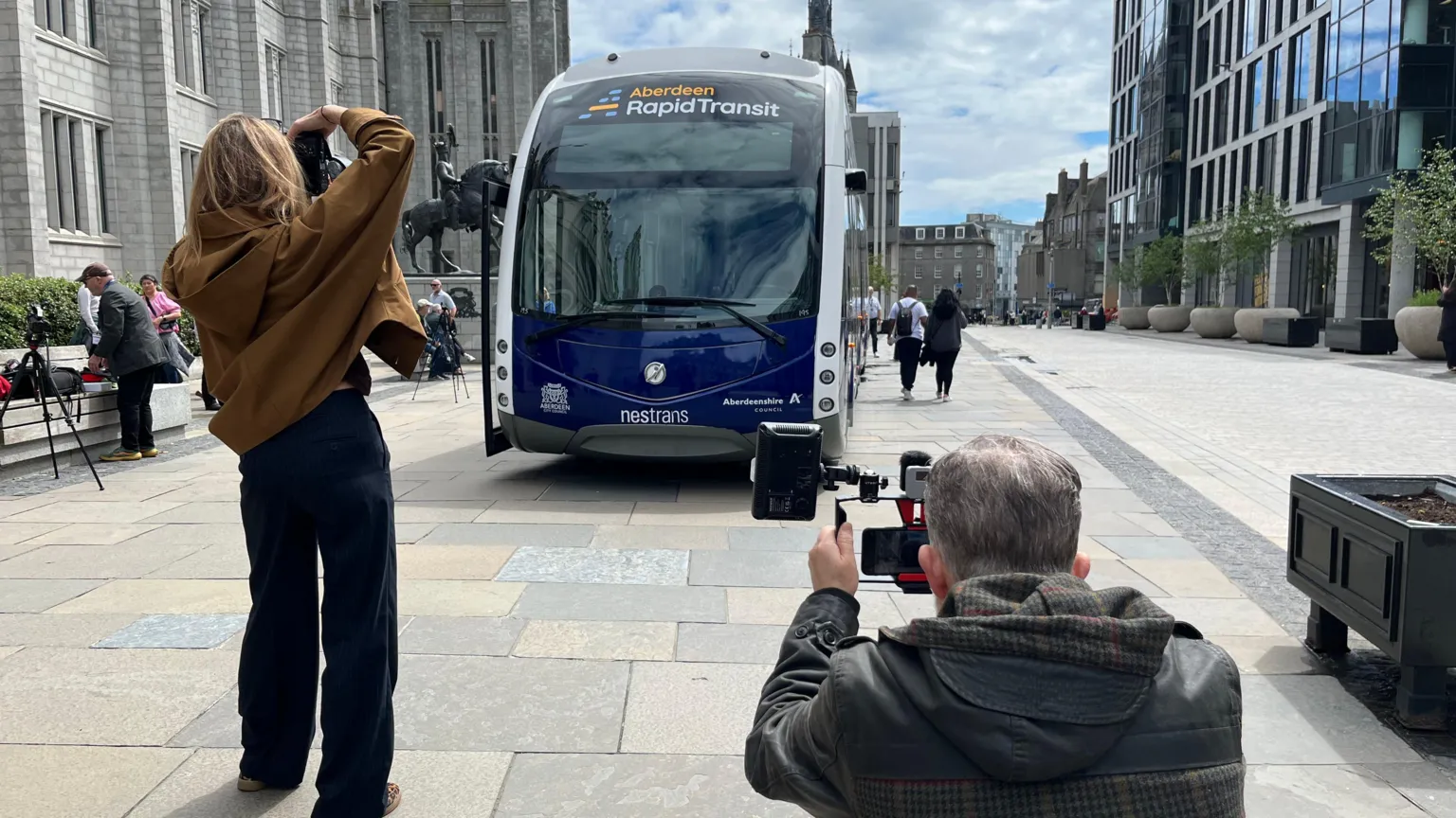 Two journalists taking photos of the vehicle which is parked on a pavement. 