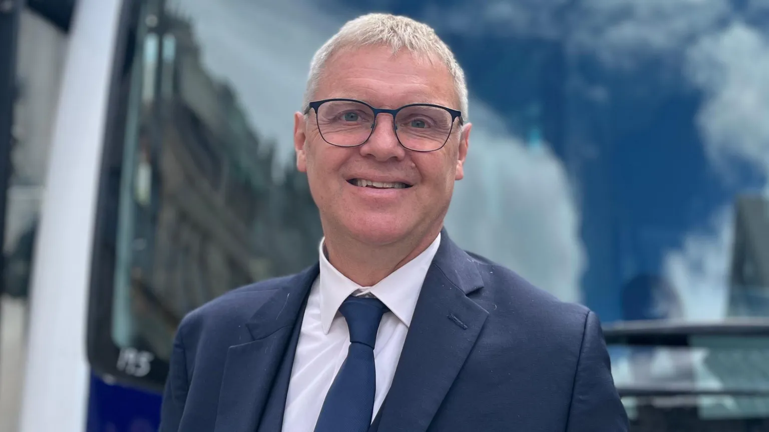 Paul Finch stood in front of the bus and smiling at the camera. He is wearing a suit and tie and glasses. 