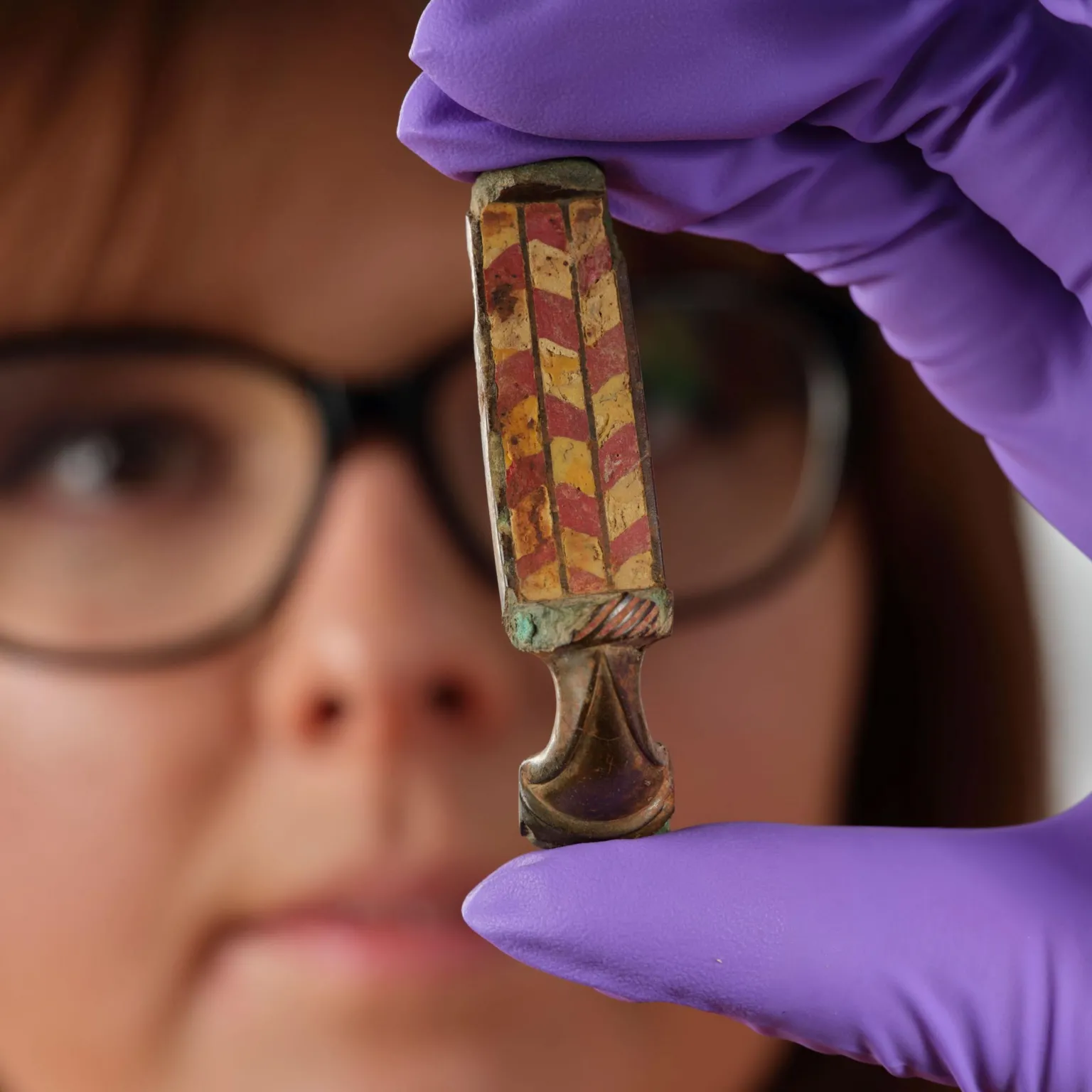  A woman wearing glasses holds up the brooch in a hand while wearing purple, protective gloves. The brooch is a a rectangle shaped object with a brown taper shaped end. The rectangle shaped section is decorated with a red and yellow chevron-like pattern.